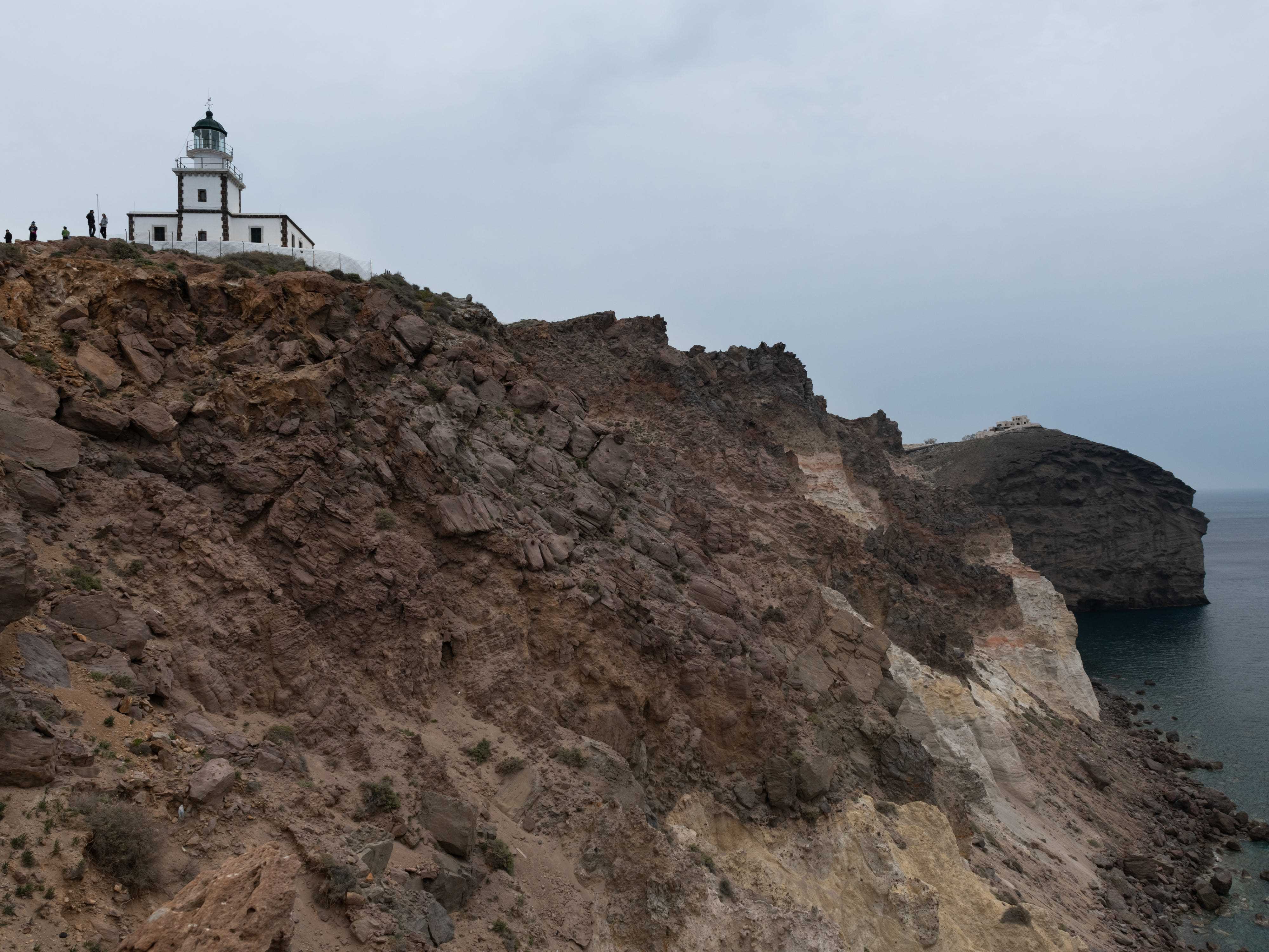 Braune Felsklippen mit Blick auf das Meer an einem düsteren Tag.  In der Ferne ein weißer Leuchtturm mit Silhouetten von Menschen