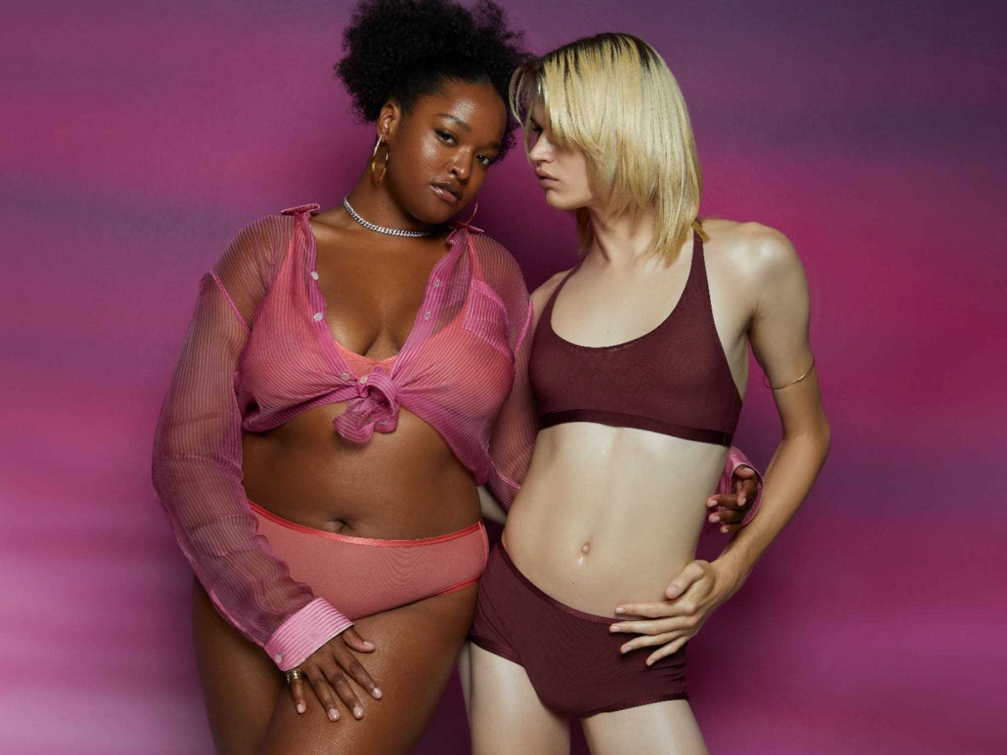 Zwei Models stehen vor einem violetten Hintergrund in rosa und kastanienbraunen Parade-Dessous.