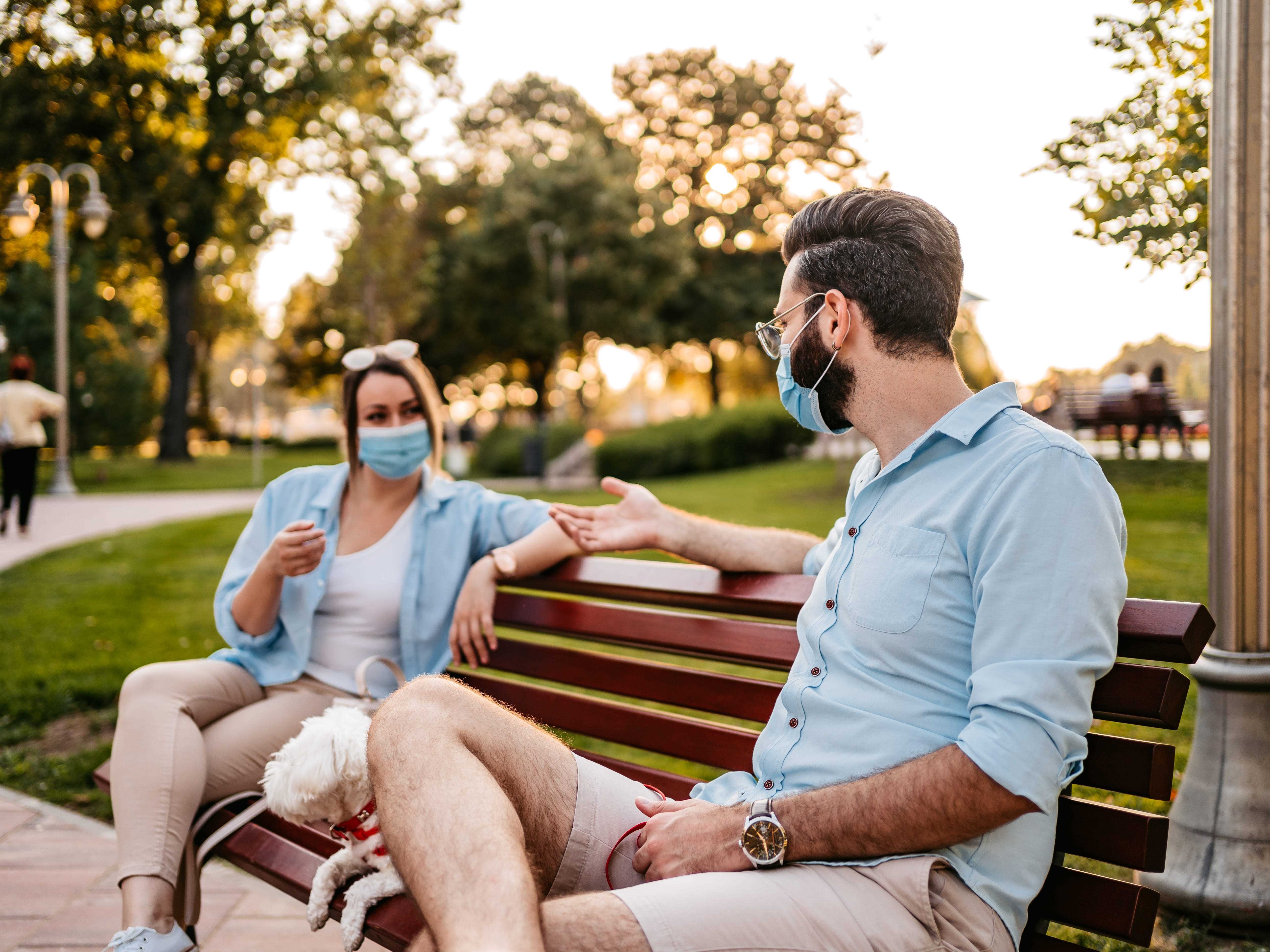 Junge süße heterosexuelle Paare, die auf einer Parkbank soziale Distanz halten und schützende Gesichtsmasken tragen.  Dazwischen sitzt ein kleiner Hund.