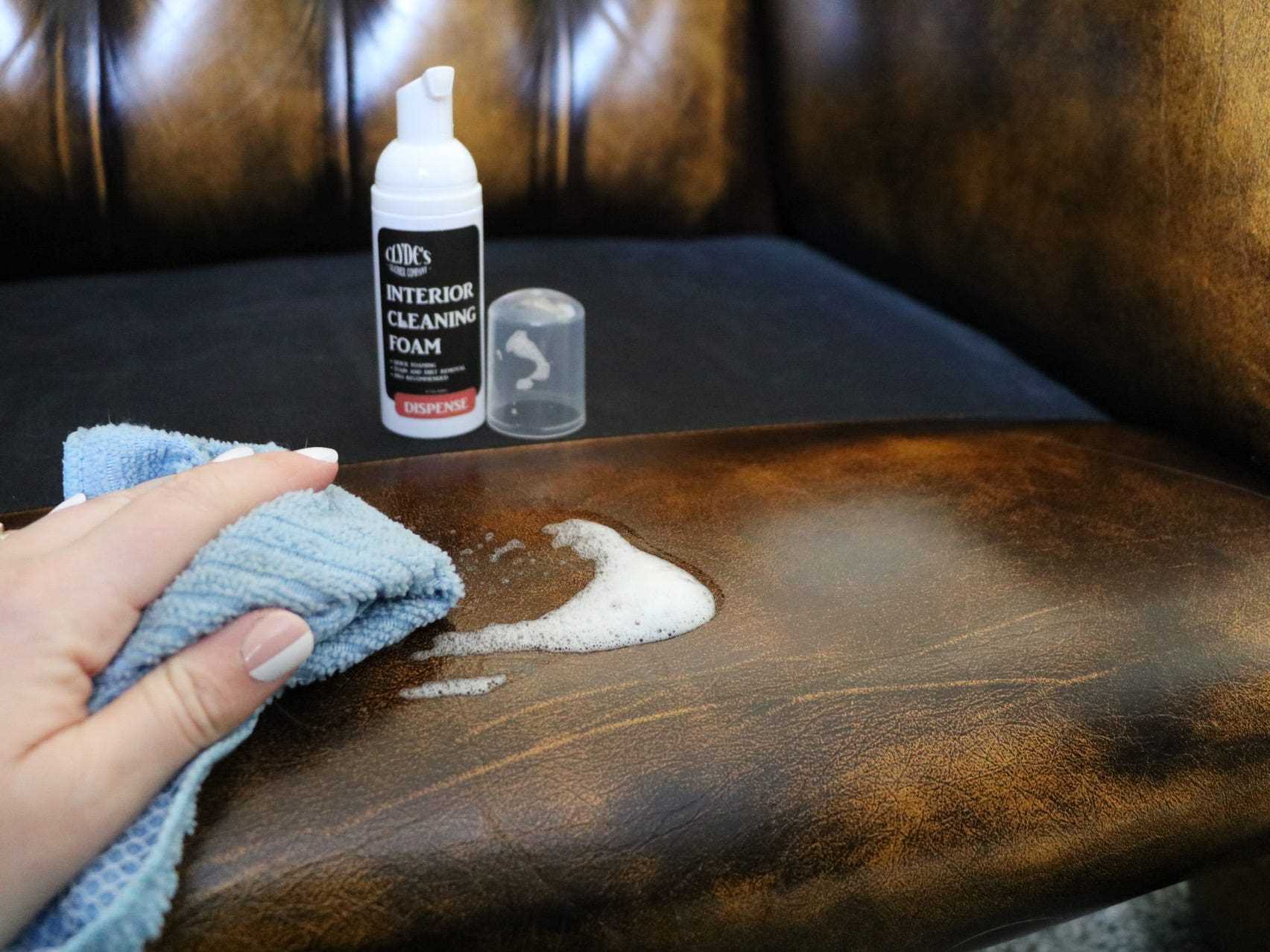 Eine Person, die den Clyde's Leather Interior Cleaning Foam mit einem Tuch auf die Armlehne eines Ledersessels aufträgt.