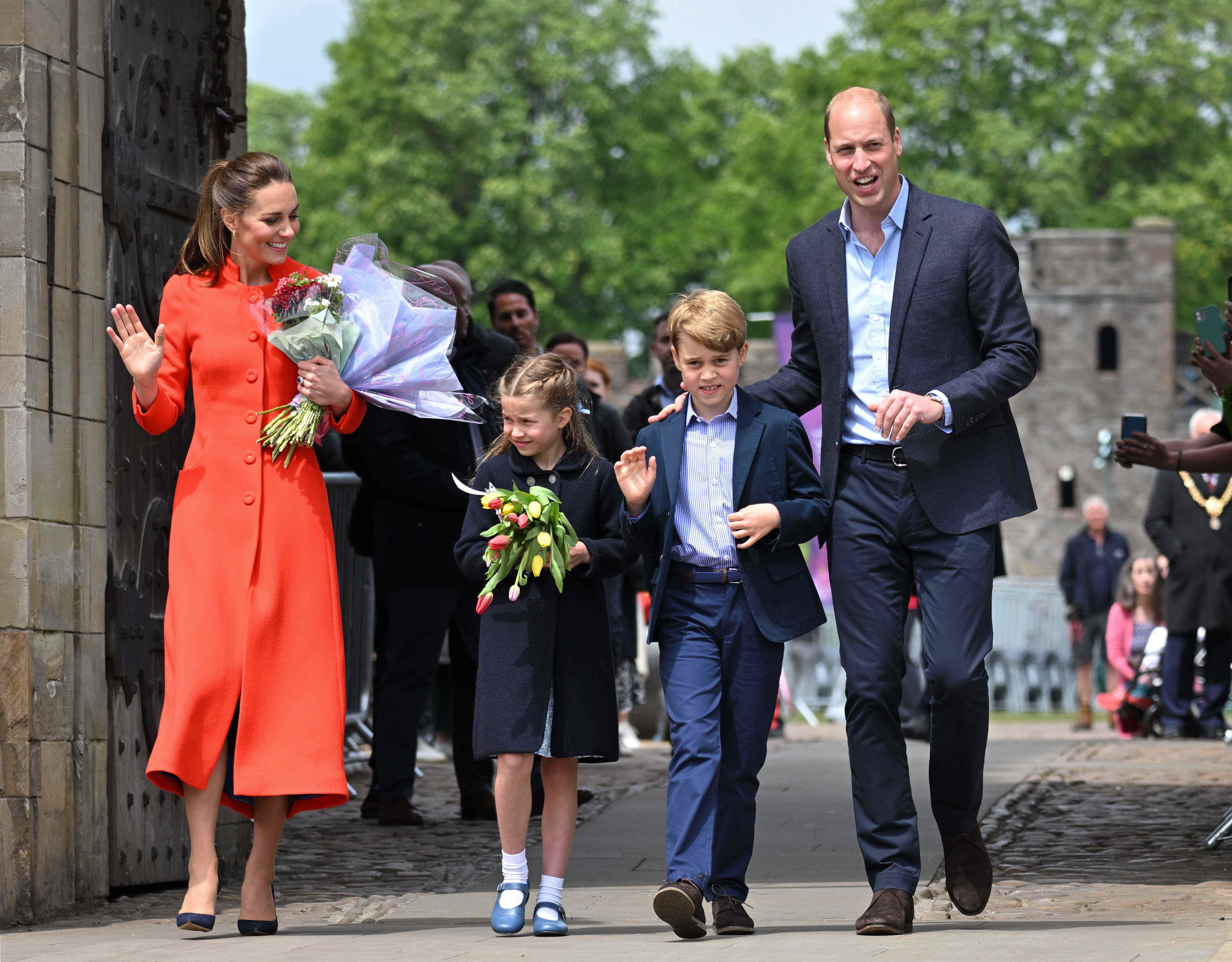 Kate Middleton in einem roten Mantel neben ihren Kindern in Marineblau und Prinz William, ebenfalls in Marineblau