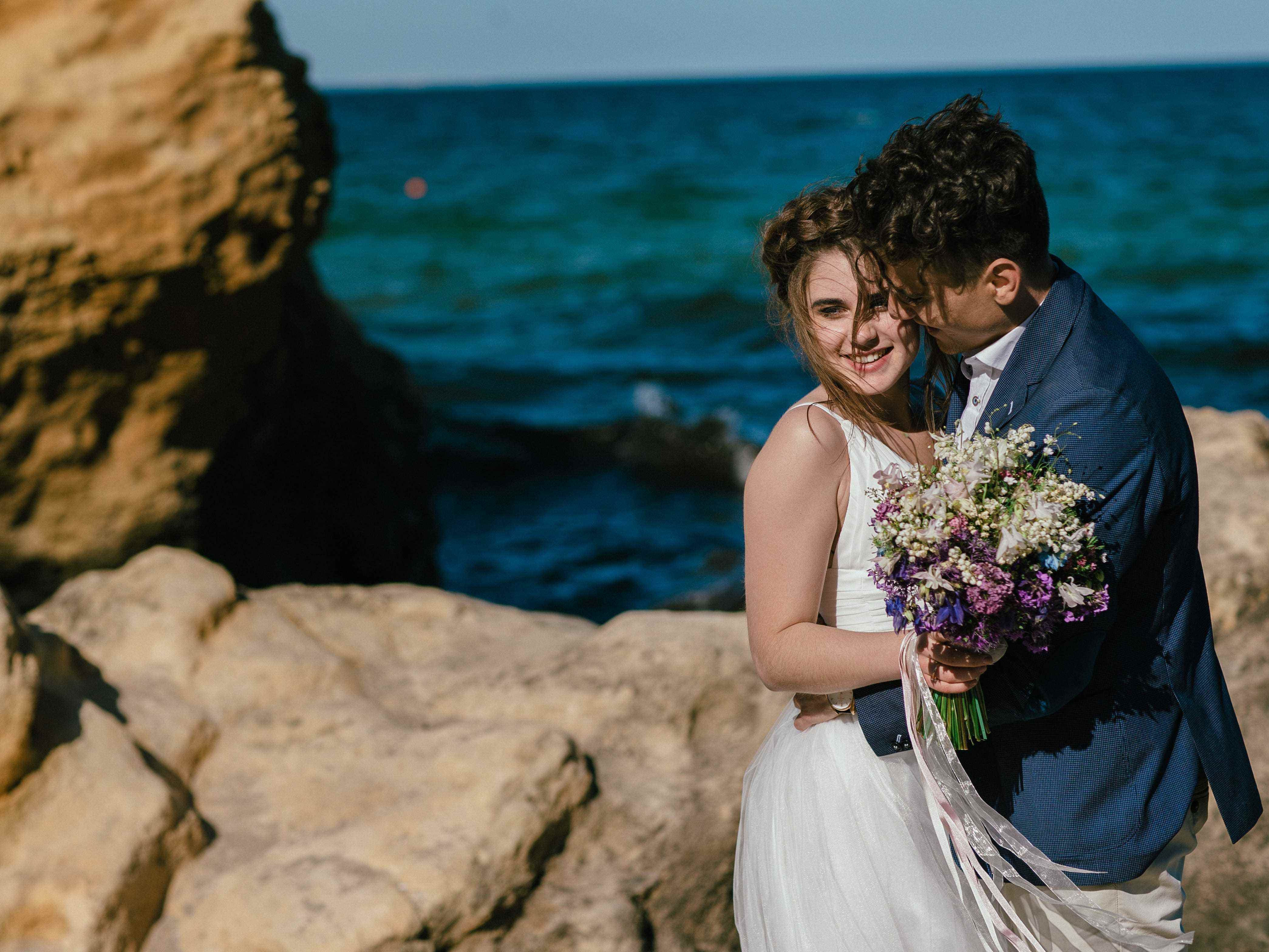 eine Braut und ein Bräutigam, die Blumen auf Felsen durch einen Strand halten