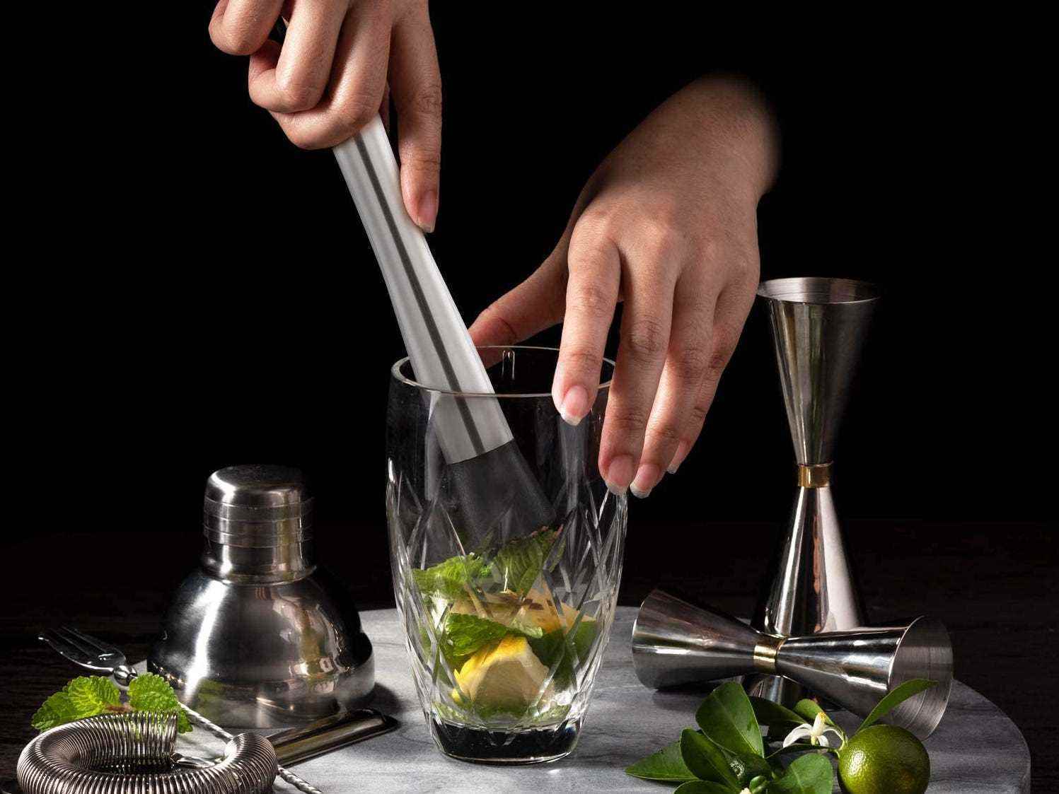 Zwei Hände mischen ein Glas Limette und Kräuter inmitten eines Cocktailsets mit losen Zutaten