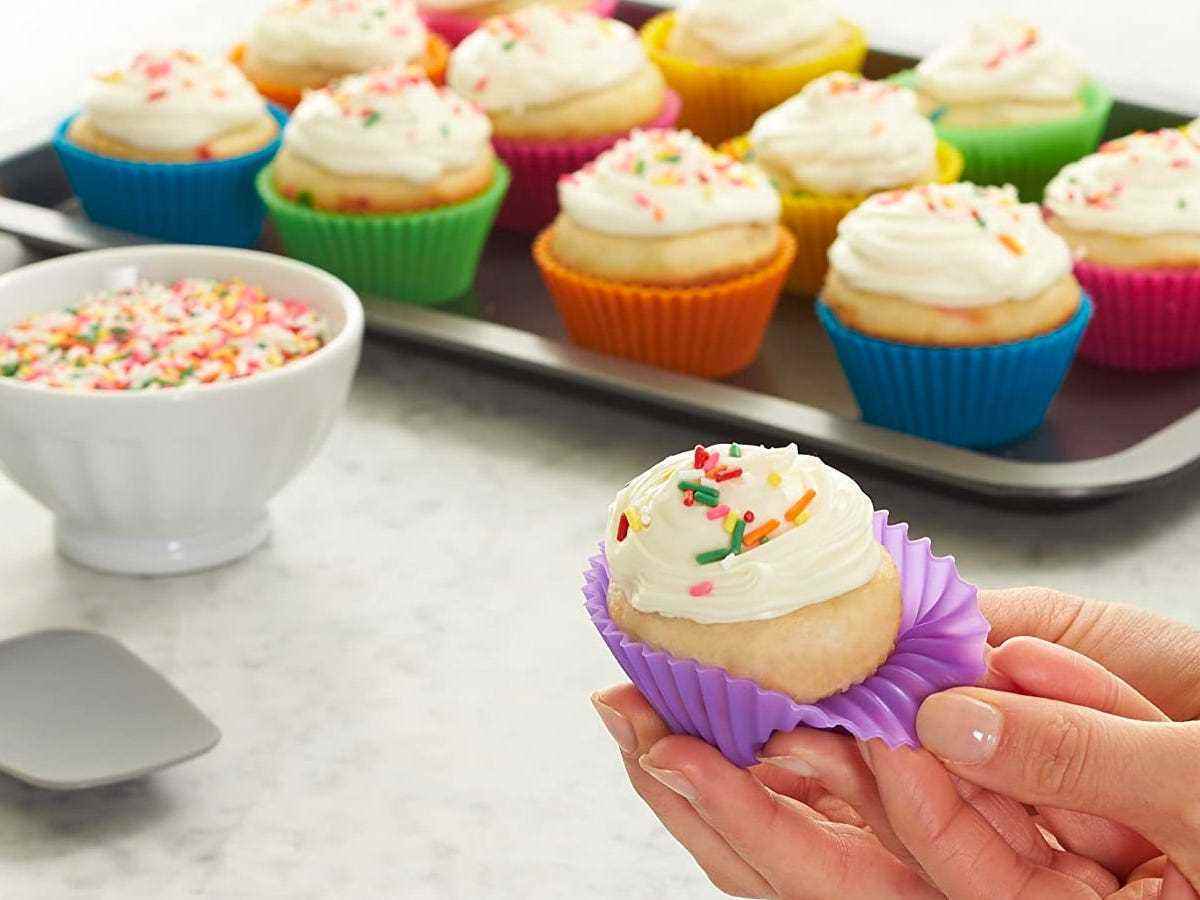 Cupcakes mit Vanille-Frost in Regenbogensilikoneinsätzen neben einer Schüssel mit Regenbogenstreuseln und einem Henkel, der einen Cupcake in einem violetten Einsatz hält