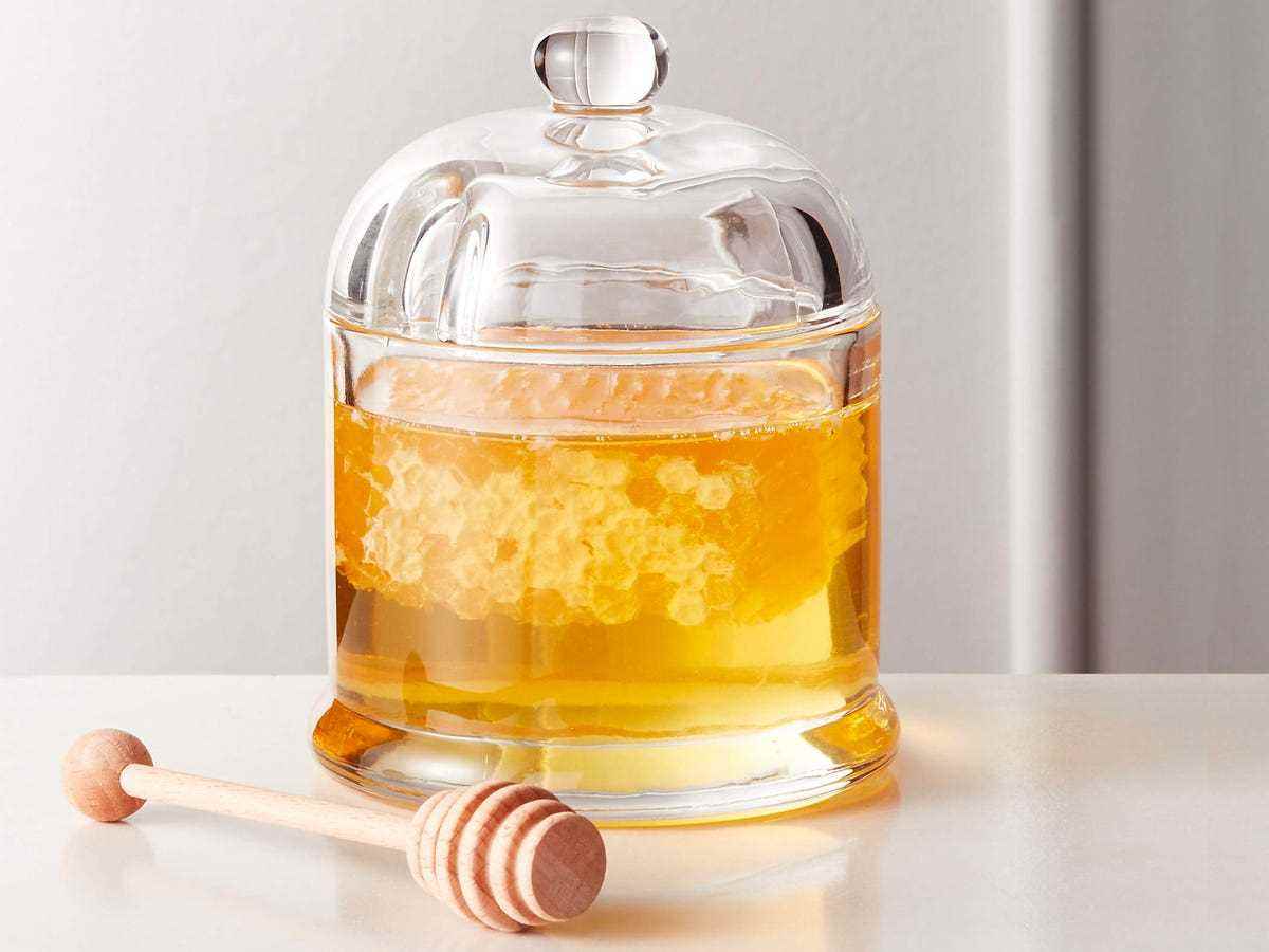 Ein durchsichtiges Glas mit abgerundetem Deckel, gefüllt mit Honig und Wabenstücken, neben einem Honigsprühstab