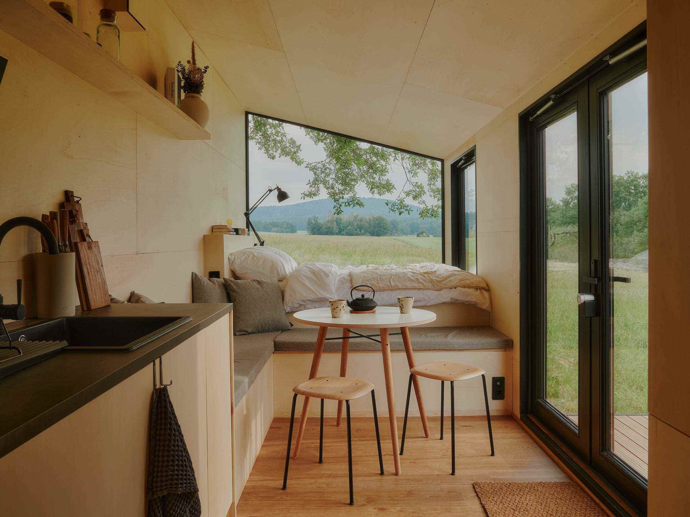 Blick in eine Raus-Hütte mit Bett, Esstisch, Küche, Fenster mit Blick in die Natur.