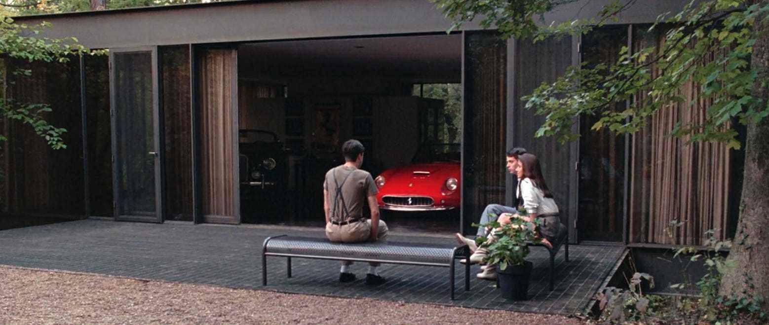 Drei Teenager sitzen auf Bänken vor einer Garage, in der ein knallroter Ferrari steht.  Das Haus ist ein Designerhaus im Highland Park mit Glastüren, die sich in die Showroom-Garage öffnen