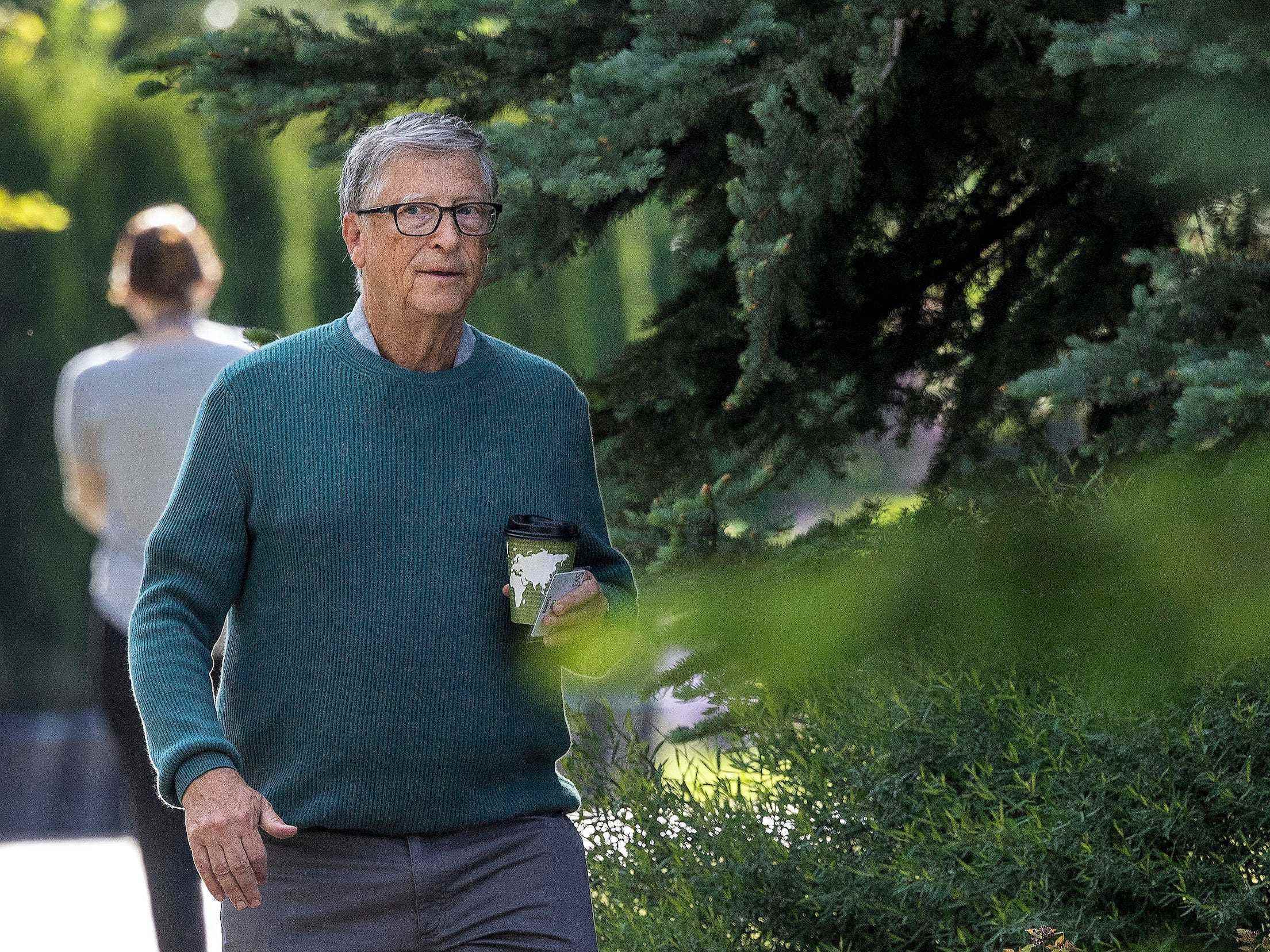 Bill Gates mit einer Kaffeetasse, die auf der Sun Valley-Konferenz zwischen Pinien spazieren geht