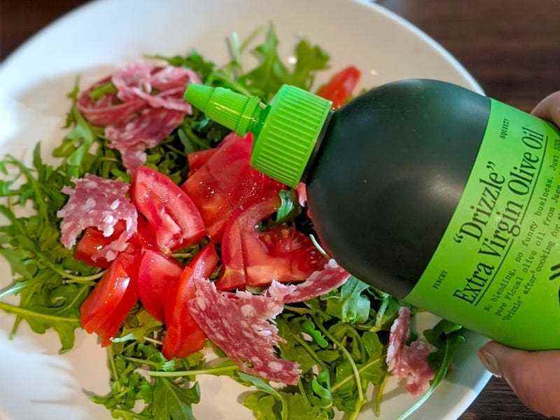 Eine grüne Flasche Graza-Olivenöl schwebt über einem Teller Rucola-Salat.