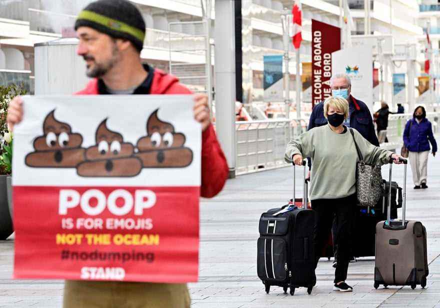 Ein Mann steht vor einem Kreuzfahrtschiff und hält ein Schild mit der Aufschrift „Poop is for emojis, not the oceans“.  Passagiere mit Gepäck sind hinter ihm zu sehen.
