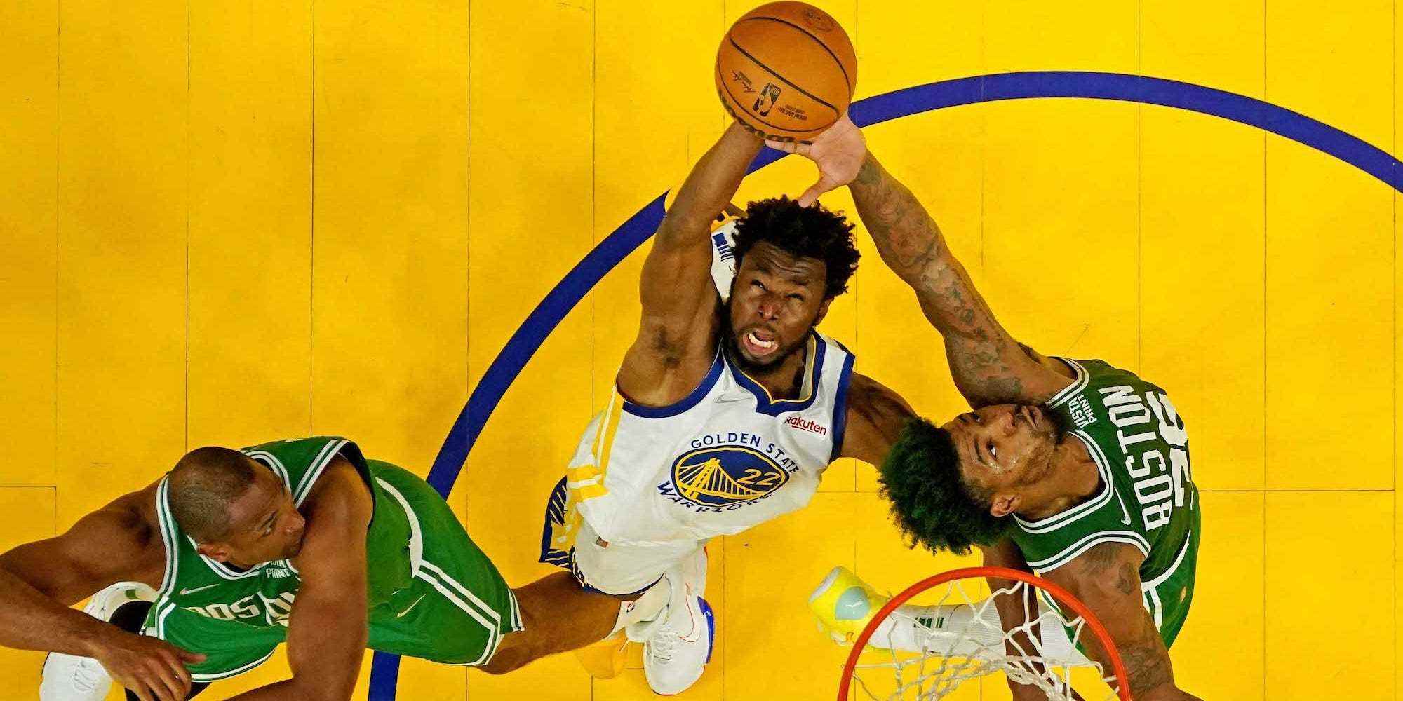 Eine Aufnahme von oben zeigt Andrew Wiggins, der in die Luft springt und einen Rebound erzielt, während er von Celtics-Spielern umgeben ist.