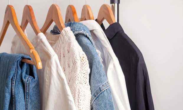Eine Auswahl an Kleidungsstücken, darunter einige Jeans, weiße Oberteile, eine Jeansjacke und ein dunkles Hemd, die an hölzernen Kleiderbügeln auf einem Gestell hängen