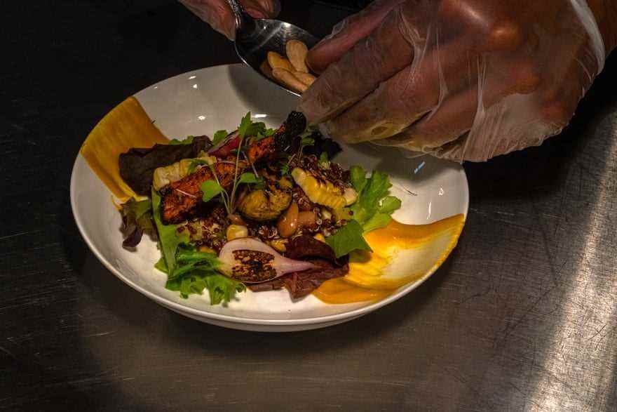 Eine Nahaufnahmeansicht eines Tellers des Essens mit Blattsalat und Mais.  Behandschuhte Hände verleihen dem Teller den letzten Schliff.