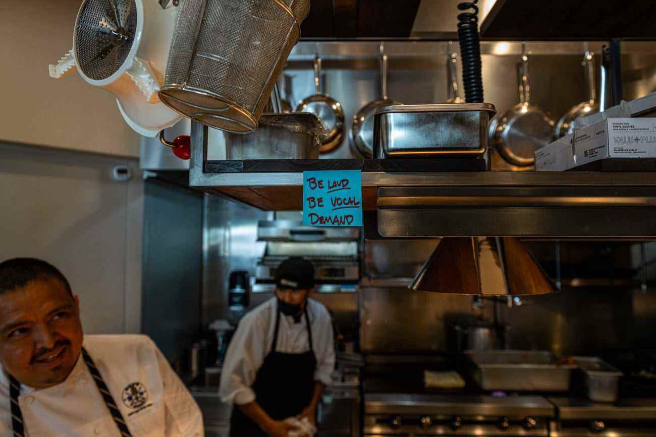 Zwei Männer arbeiten in einer professionellen Küche in der Nähe von Öfen.  Auf einem blauen Haftnotizzettel, der an einem mit glänzenden Metallwerkzeugen gefüllten Regal befestigt ist, steht: „Sei laut, sei laut, fordere!“