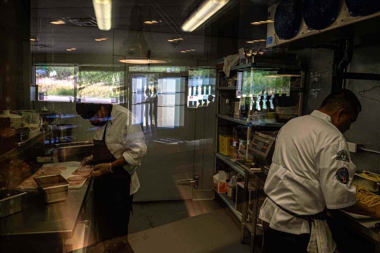 Durch ein Fenster sieht man zwei Männer, die mit Essen arbeiten, während sich der Rest der Küche im Glas spiegelt.