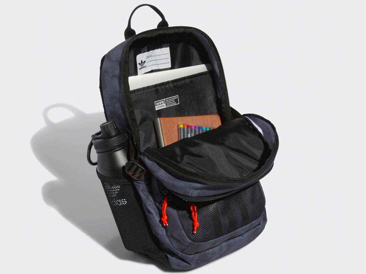 Ein Adidas Energy Rucksack, teilweise geöffnet, um Notizbücher, Stifte und andere Schulmaterialien vor einem grauen Hintergrund zu zeigen