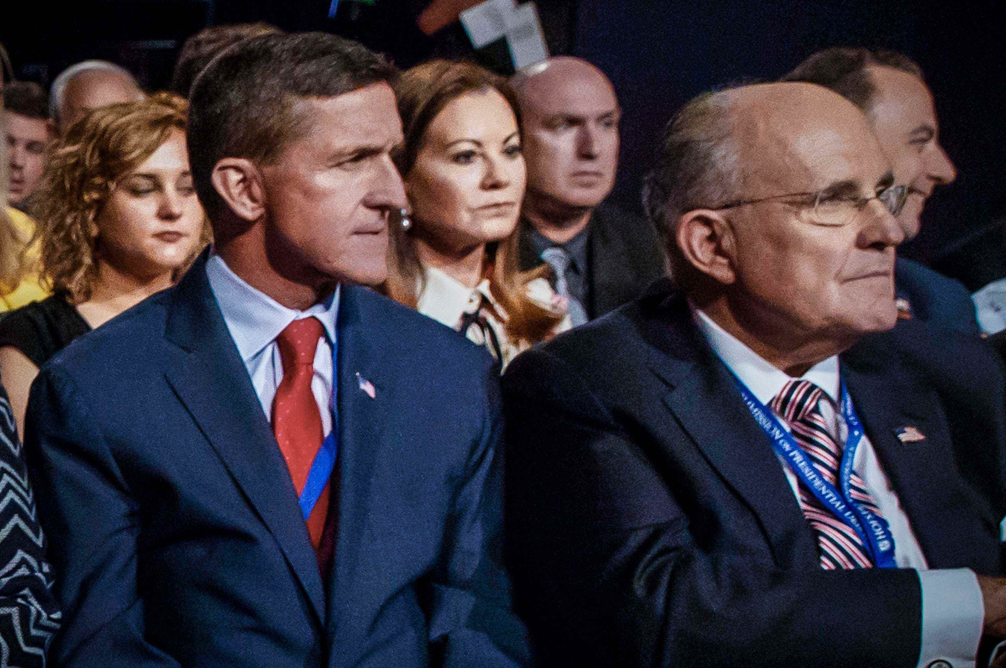 Links: Michael Flynn sitzt in einem blauen Anzug und einer rot-blauen Krawatte in einer Menschenmenge;  Rechts: Rudy Giuliani sitzt neben ihm in einer rot-schwarz gestreiften Krawatte und einem schwarzen Anzug