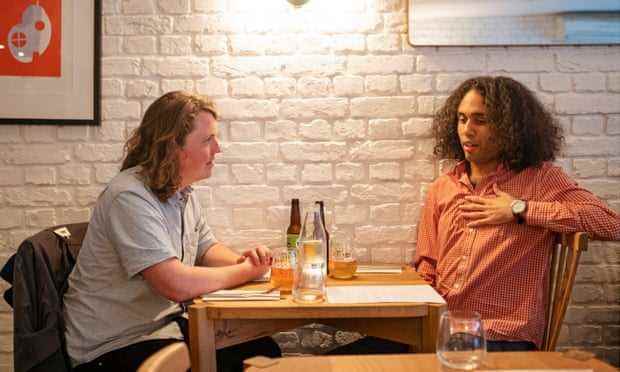 Jasper und Miles sitzen an einem Restauranttisch vor einer weiß gestrichenen Ziegelwand