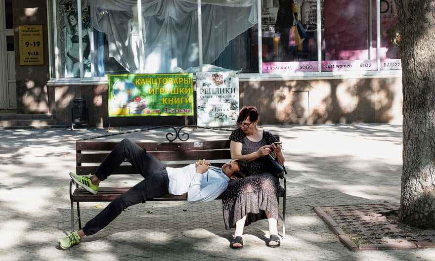 Ein junger Mann in gelben Turnschuhen liegt auf einer Bank unter einem Baum, den Kopf auf dem Schoß einer älteren Frau, während sie durch ihr Telefon scrollt.