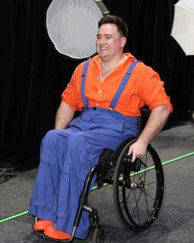Ein Mann trägt ein orangefarbenes Hemd und Schuhe und einen blauen Overall, während er im Rollstuhl sitzt