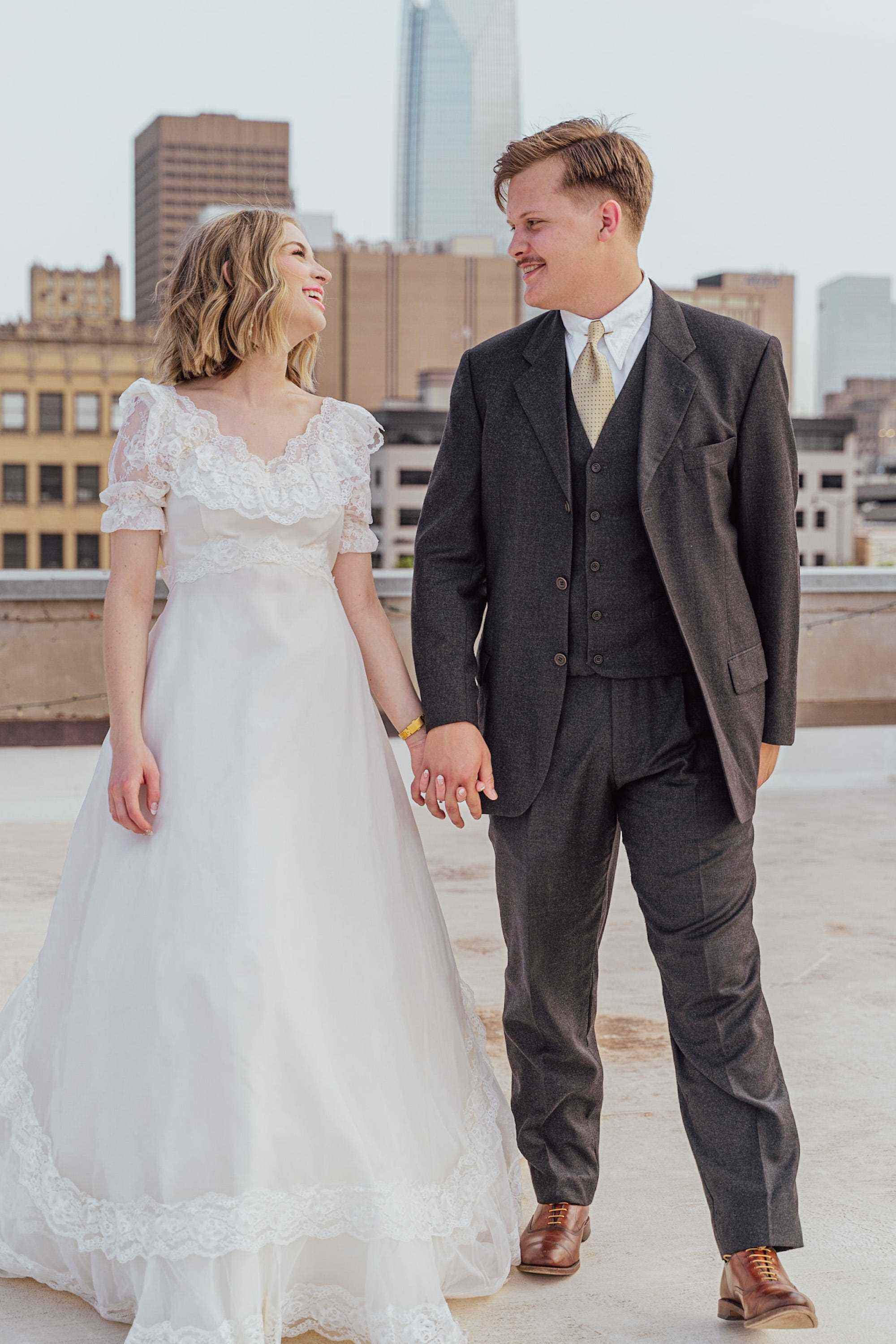 Eine Braut und ein Bräutigam halten sich in ihrer Hochzeitskleidung auf einem Dach an den Händen.