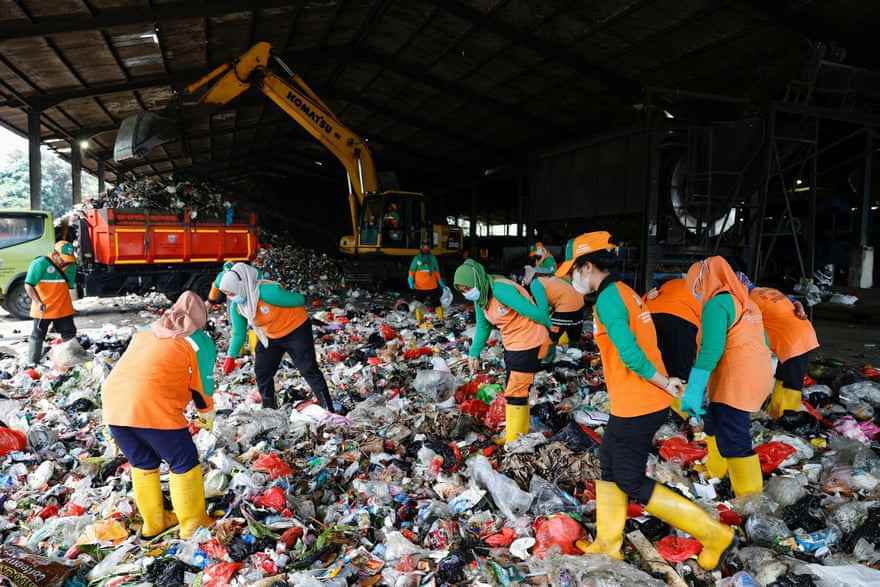 Frauen in Masken, orangefarbener Uniform und Gummistiefeln wühlen sich durch eine riesige Müllhalde