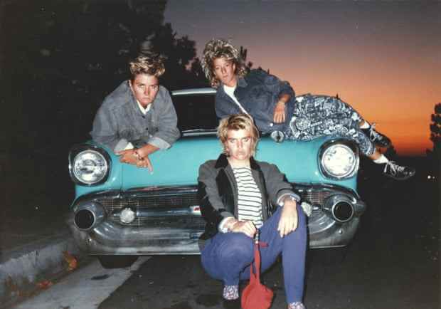 Die Surfer Jodi Cooper, Toni Sawyer und Pam Burridge sitzen in den 80er Jahren auf einem Oldtimer mit dem Sonnenuntergang im Rücken.