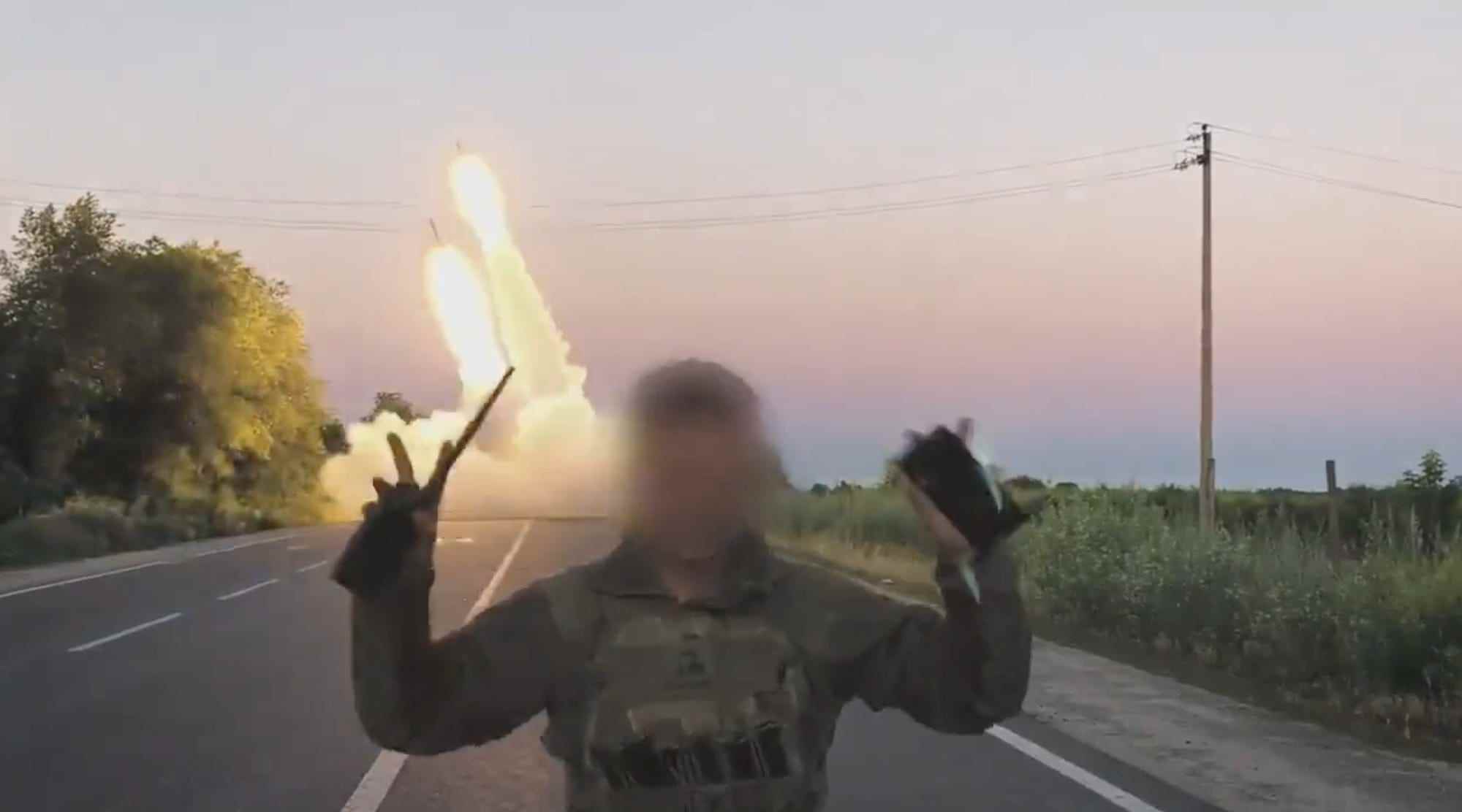 Ein Standbild aus Filmmaterial, das vom ukrainischen Verteidigungsministerium geteilt wurde, zeigt einen Soldaten, dessen Gesicht verdeckt ist, der seine Hände in einem „v für den Sieg“ hebt, während ein von den USA gespendetes HIMARS-System im Hintergrund Raketen abfeuert.
