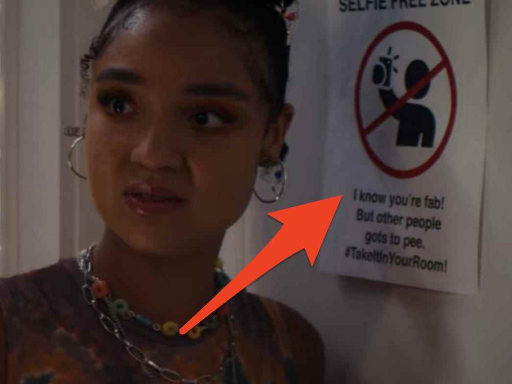 Cara steht auf einer College-Party vor einem No-Selfies-Schild an einer Badezimmertür und blickt in beide Richtungen
