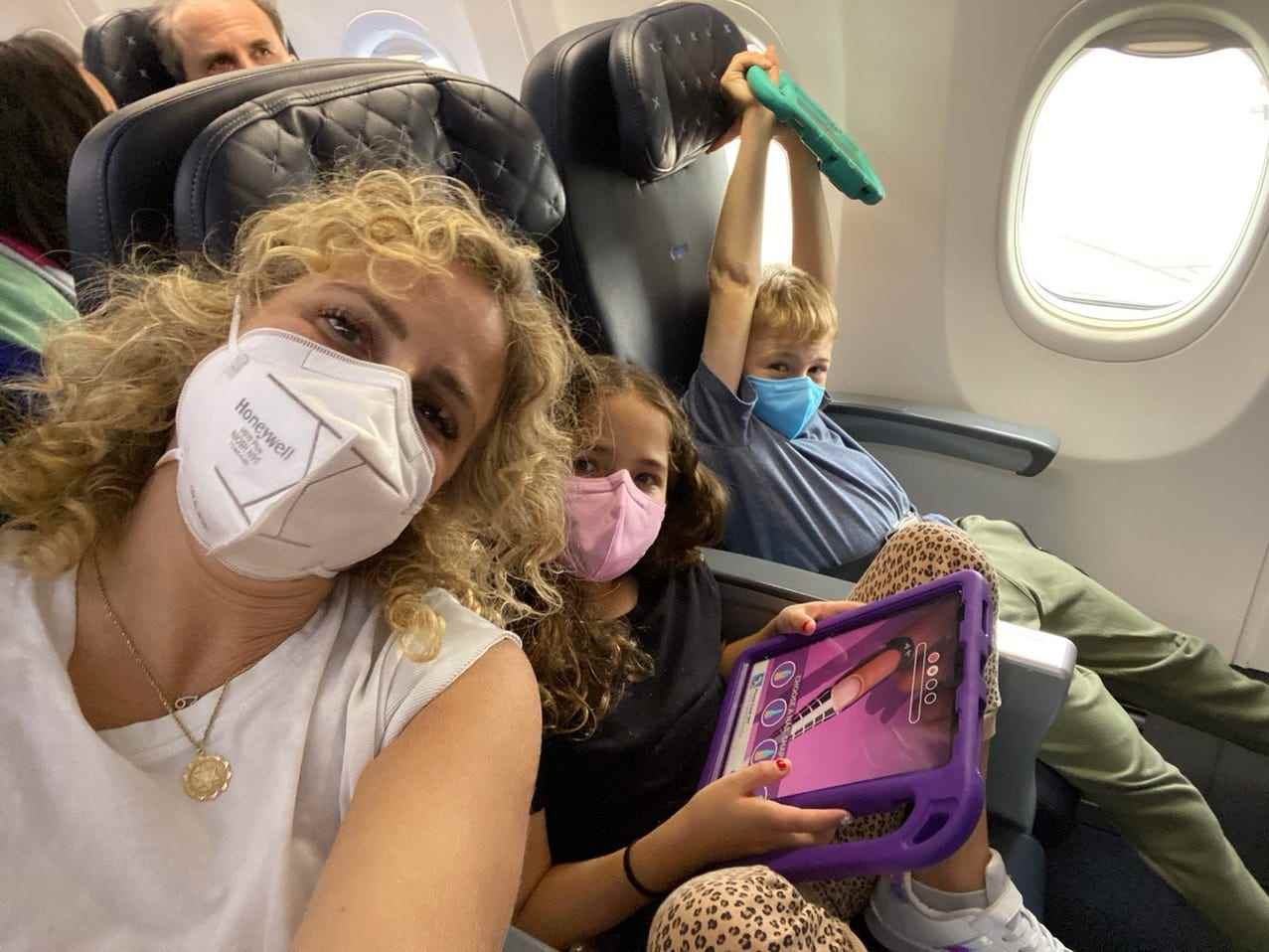 Eine Frau mit Mundschutz macht in einem Flugzeug ein Selfie mit zwei Kindern.