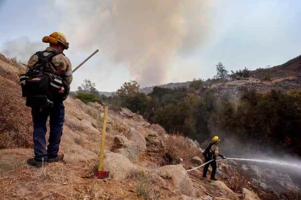 Zwei Feuerwehrleute stehen auf einem Bergrücken und zielen mit einem Feuerwehrschlauch auf eine heiße Stelle, während im Hintergrund eine Rauchwolke aufsteigt.