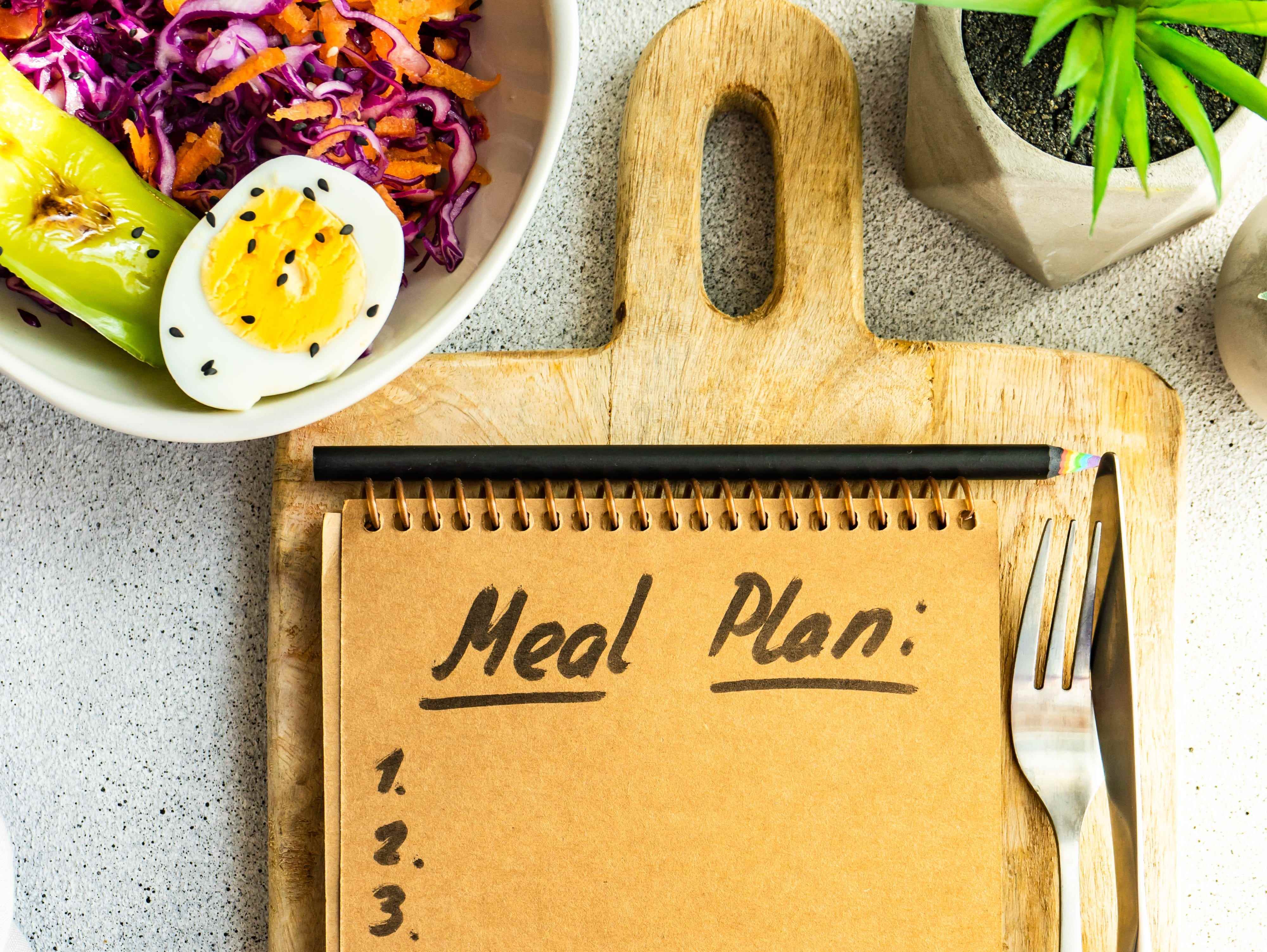 Draufsicht auf eine Schüssel mit Rotkohl, Karotten, grünem Pfeffer und Salat mit gekochten Eiern und Notizblock mit einer Essensplanliste