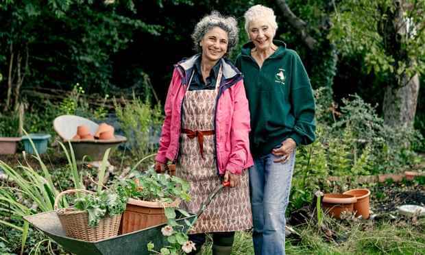 Rosa Mechoni, die Hände auf einer Schubkarre, steht neben Helen Webster in Helens Garten, beide lächeln