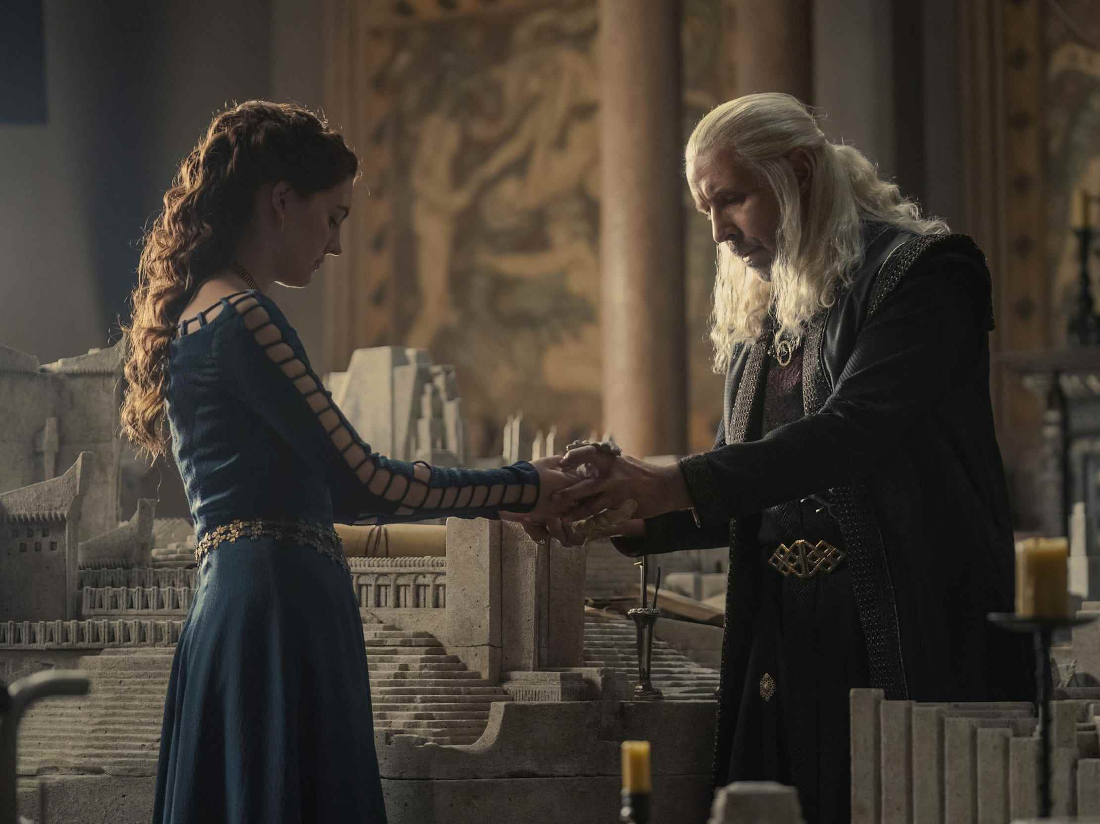 Ein junges Mädchen in einem blauen Kleid hält einem älteren Mann mit langen silbernen Haaren einen steinernen Drachen entgegen.