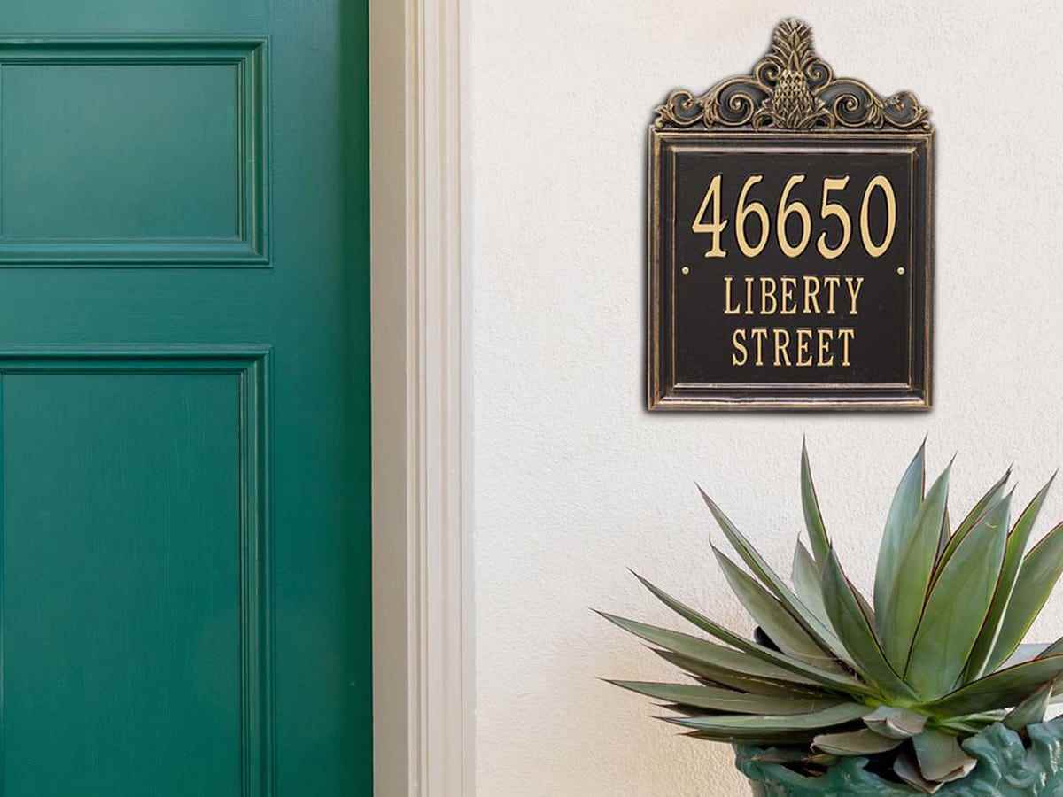 Ein dekoratives schwarzes Adressschild mit goldenem Text „46650 Liverty Street“ und goldenem Ananas-Akzent ist vor einem hellbraunen Hintergrund neben einer blaugrünen Tür angebracht.