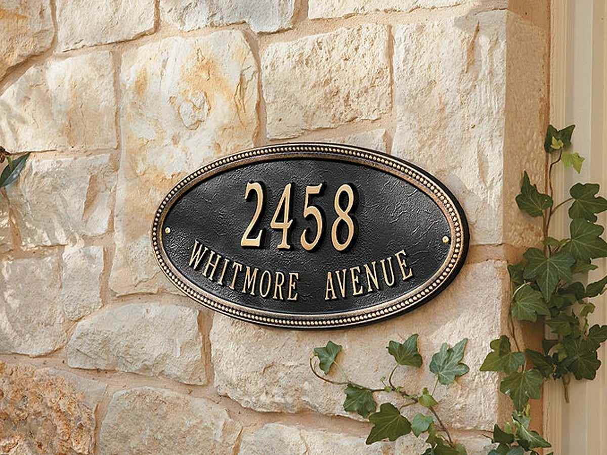 Eine ovale schwarze Plakette mit gefaltetem Text und Rand zeigt die Adresse „2458 Whitemore Avenue“ und ist an der Seite eines Steinfassadenhauses angebracht.