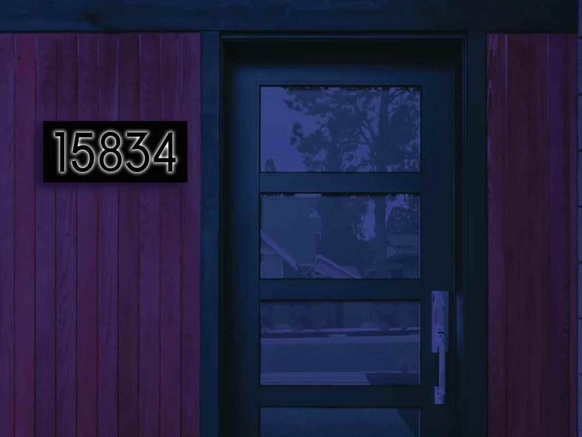 Ein rotes Brett- und Lattenhaus wird im Dunkeln mit einer schwarzen Hausnummerntafel angezeigt, die hinter den Nummern 15834 eine Hintergrundbeleuchtung hat.