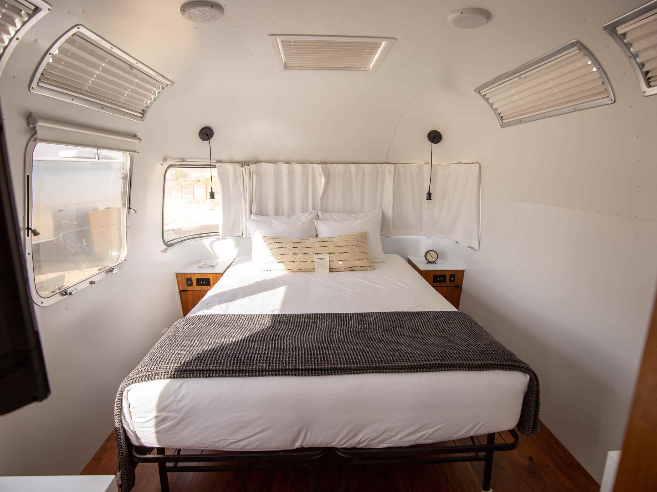 Ein Schlafzimmer im Airsteram-Wohnwagen von Autocamp Joshua Tree mit weißen Akzenten, einem Bett mit Kissen und Vorhängen über den Fenstern.