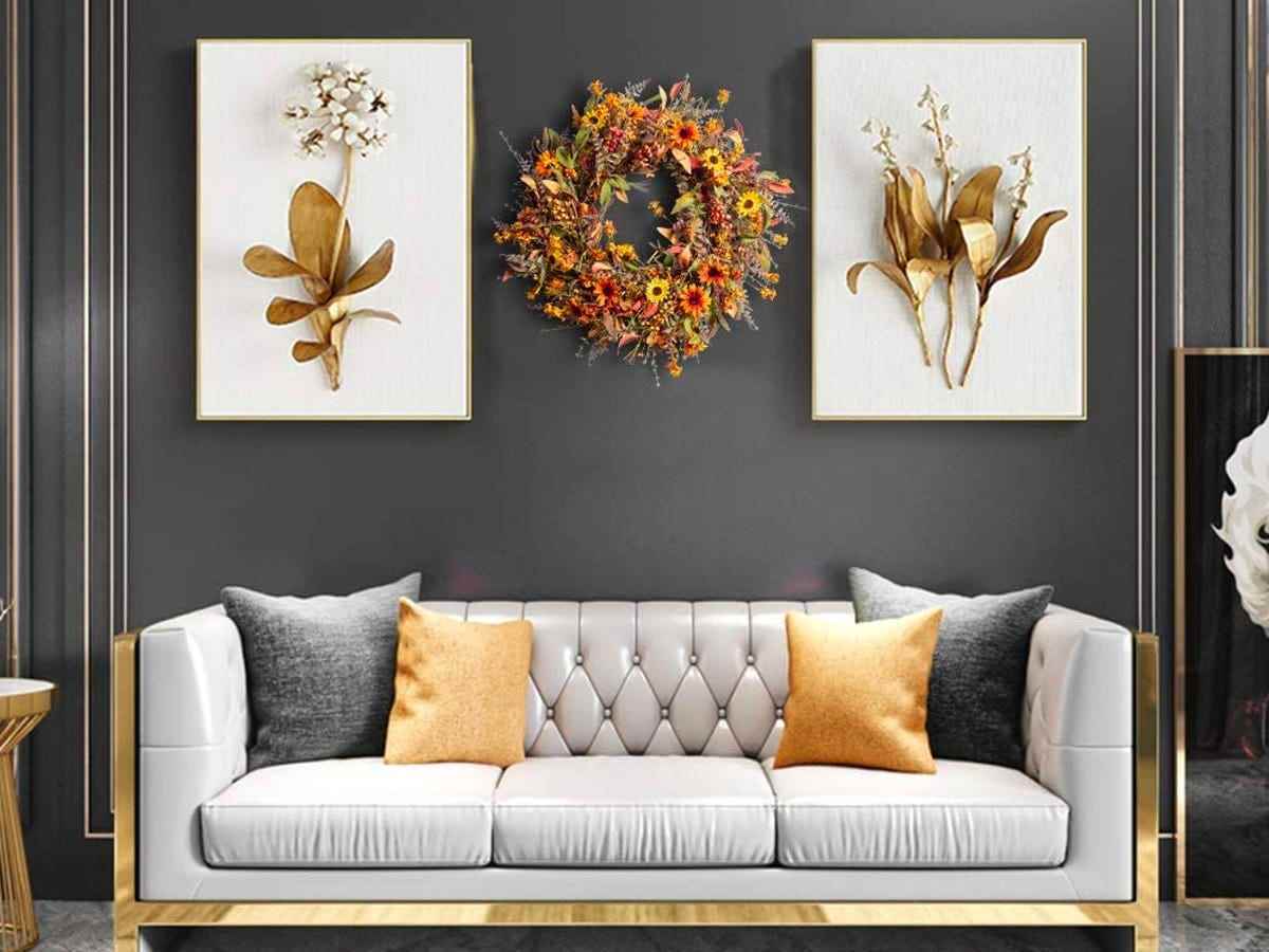 Ein Kranz mit gelben und orangefarbenen Blumen und Blättern, der zwischen zwei Gemälden an einer grauen Wand über einer hellgrauen Couch hängt