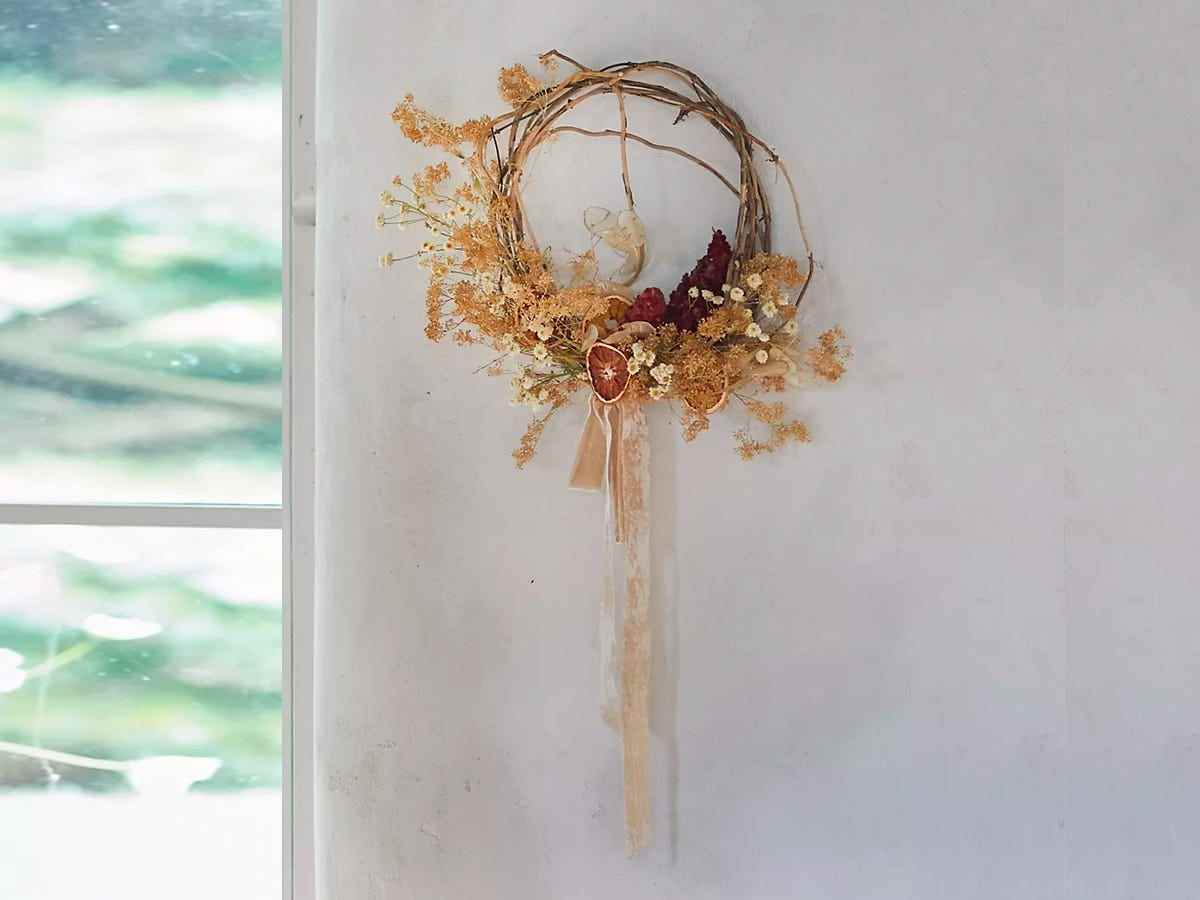An einer weißen Wand hingen eine Rebenbasis, getrocknete Blumen, Samenkapseln, getrocknete Zitrusfrüchte und ein Kranz aus Seidensamtbändern