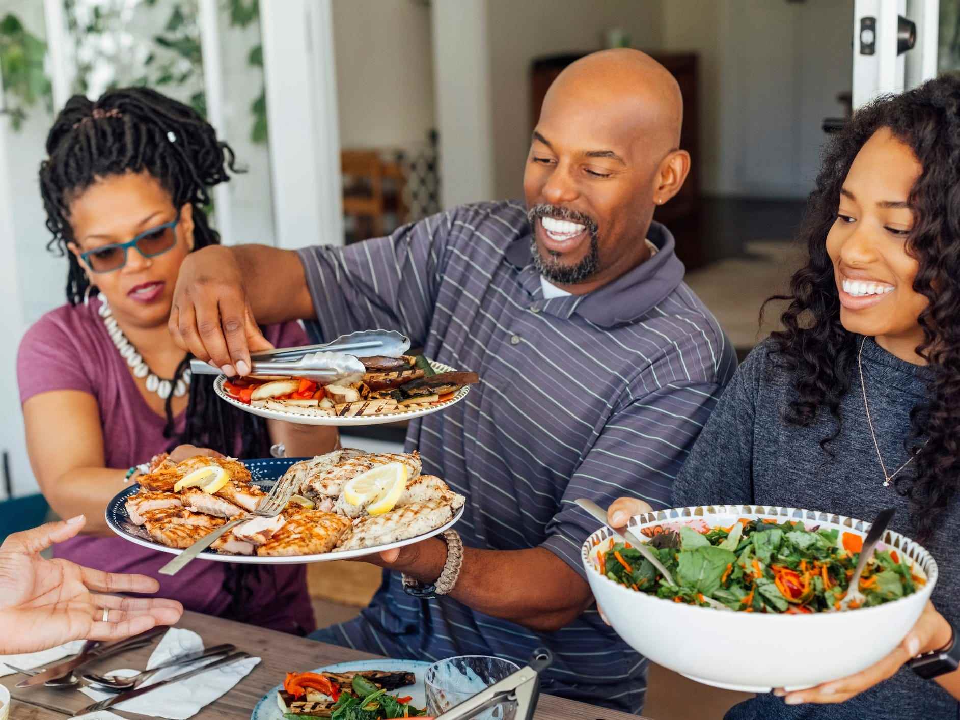 Drei lächelnde Menschen an einem Tisch im Freien, vorbei an Tellern mit Salat und Grillgerichten