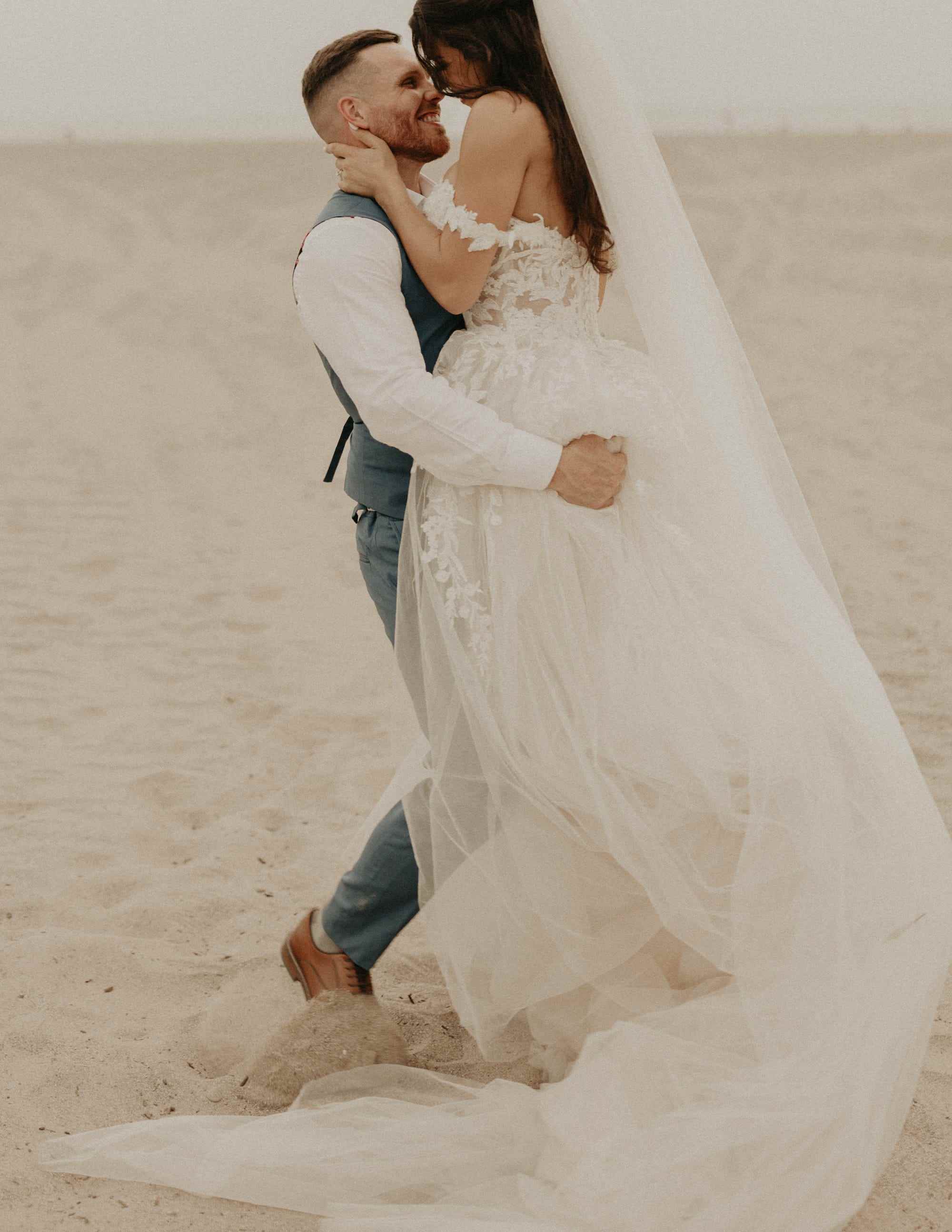 Ein Bräutigam hebt seine Braut an einem Strand hoch und sie lehnen ihre Köpfe aneinander.