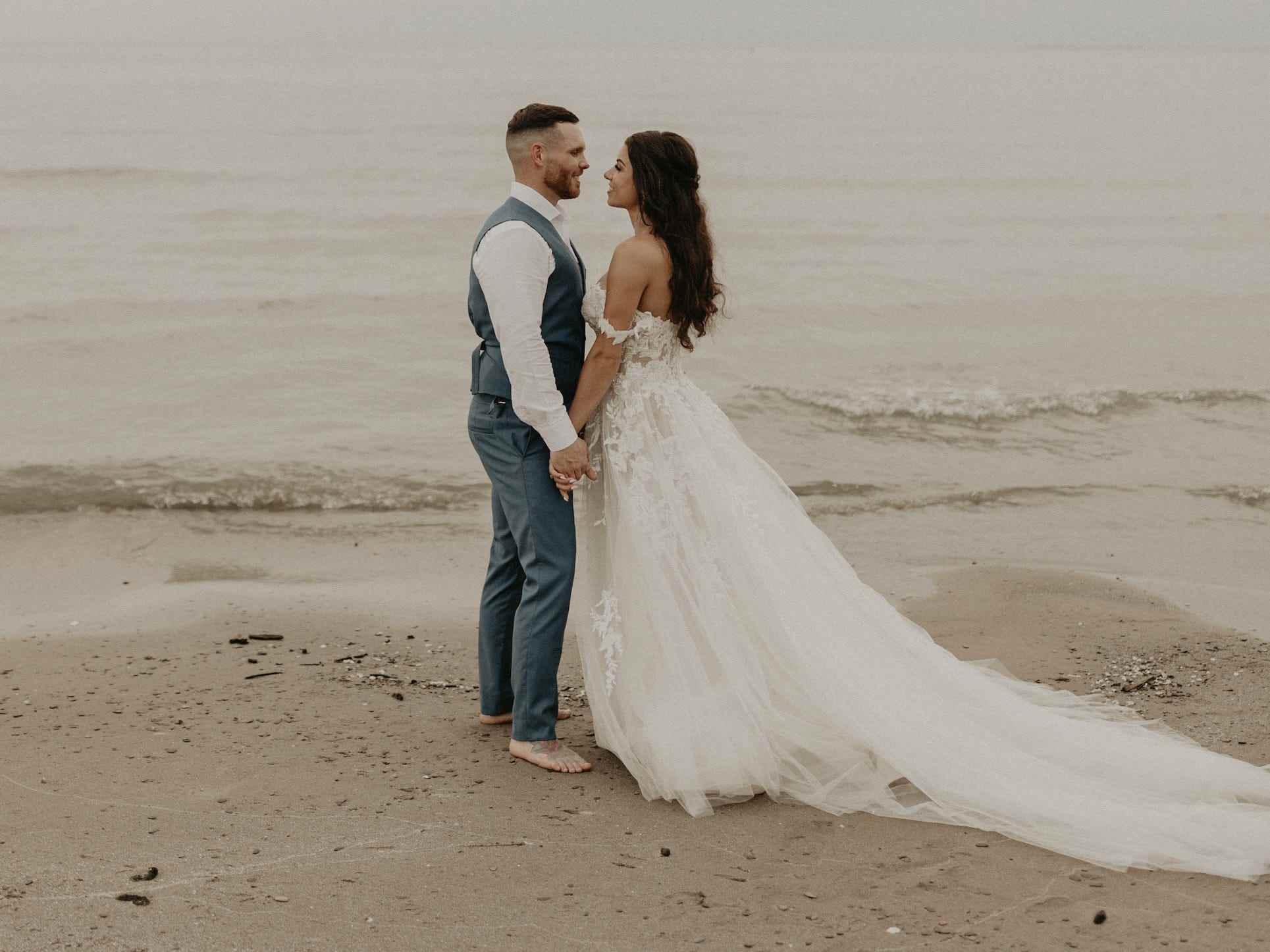 Eine Braut und ein Bräutigam halten Händchen und schauen sich vor dem Ozean an.