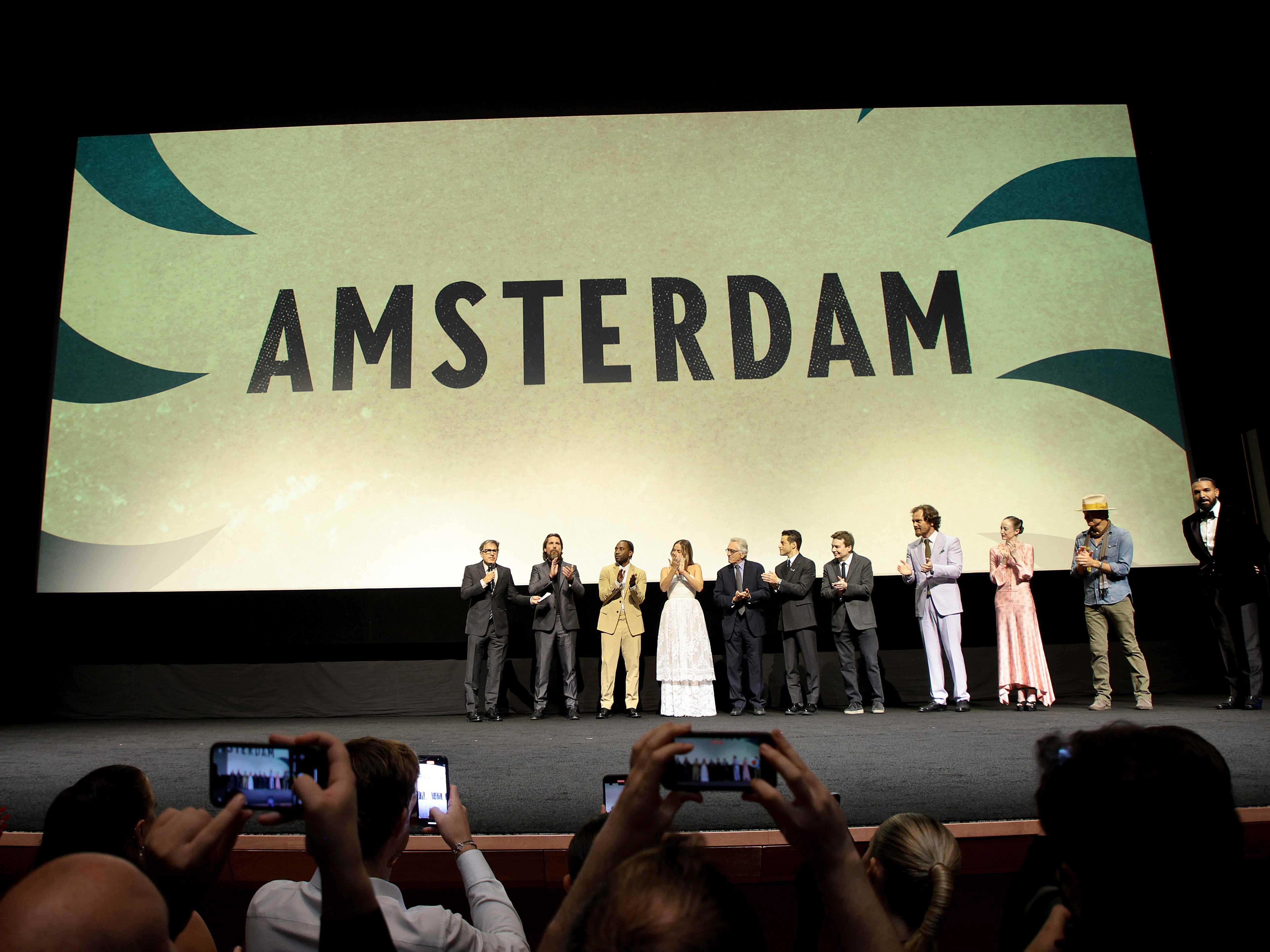 Die Darsteller von „Amsterdam“ stehen vor einer großen Leinwand mit dem Namen des Films, während die Zuschauer mit ihren Handys fotografieren