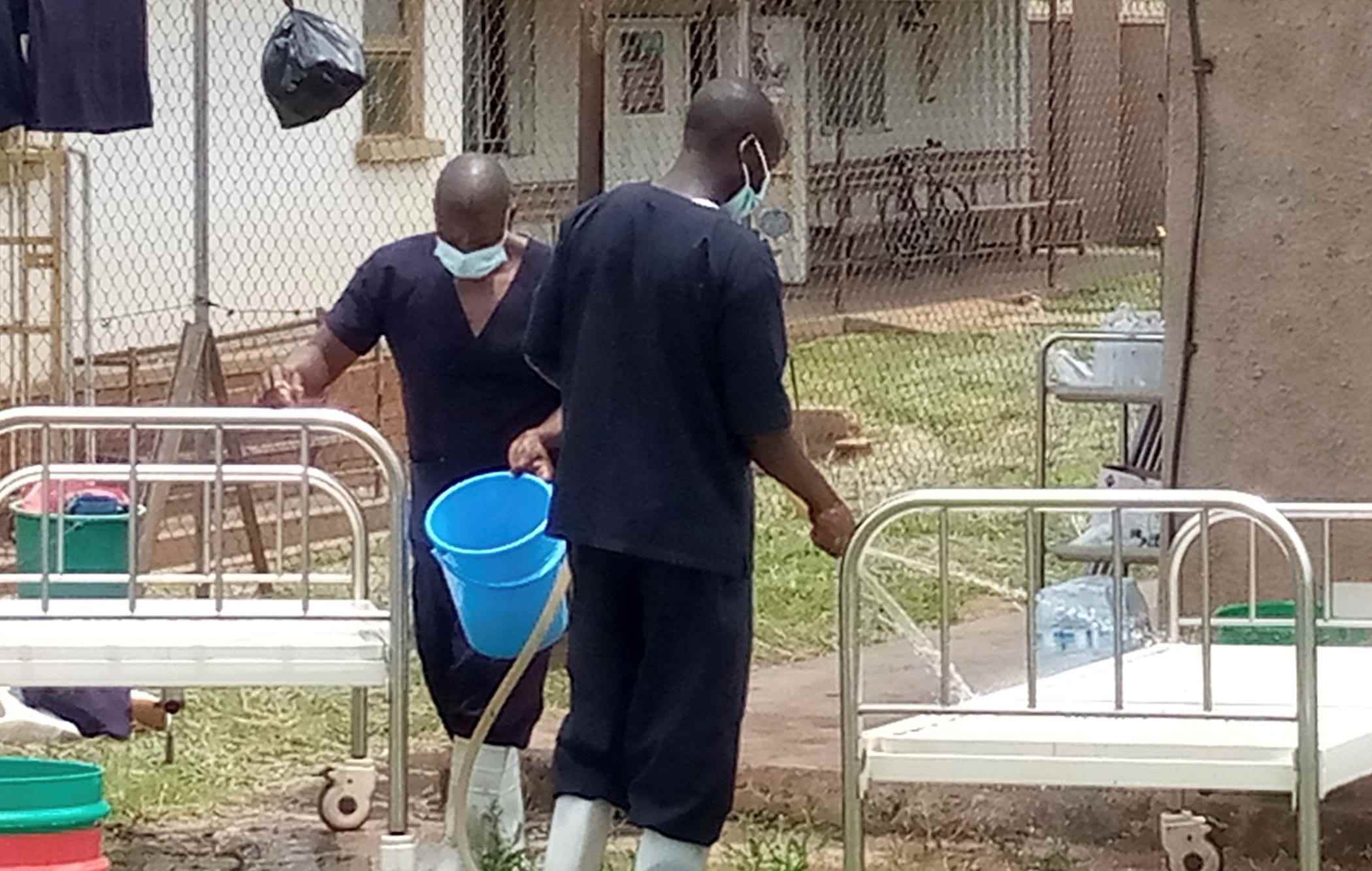 Zwei Männer in Kitteln und Masken draußen, die Betten von Ebola-Patienten mit Eimern desinfizieren