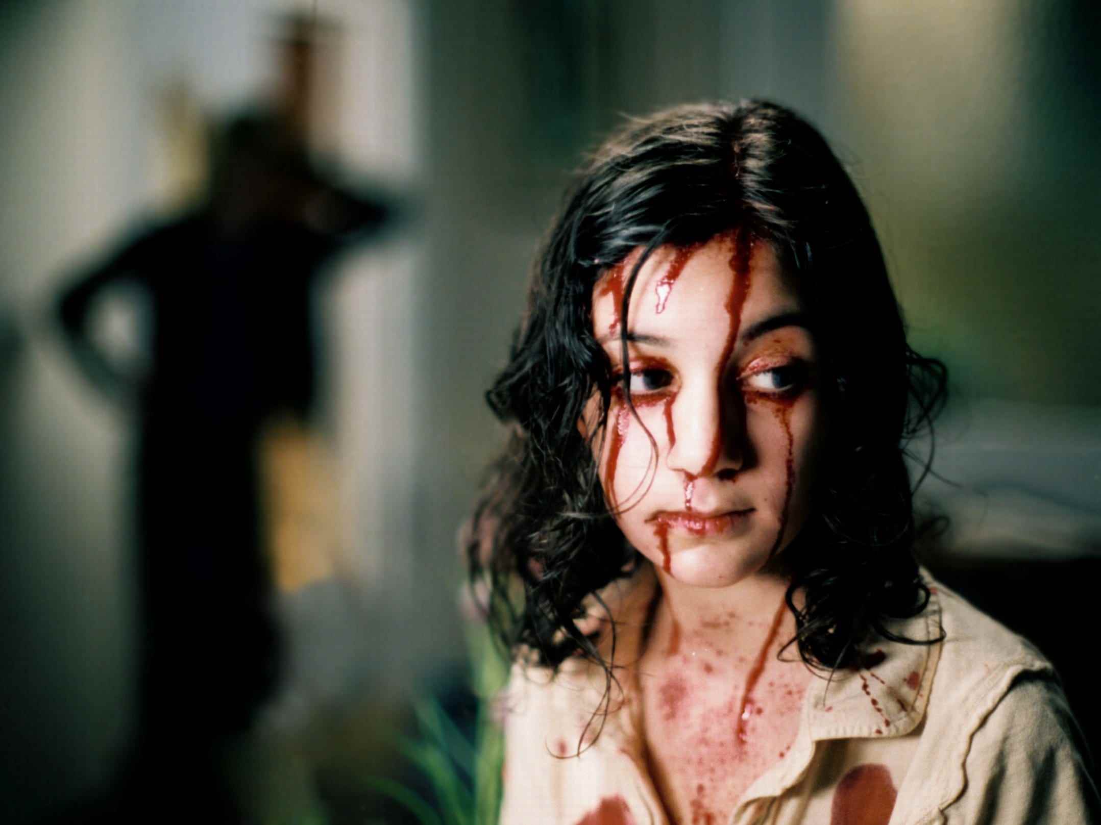 Ein Standbild aus dem Horrorfilm „Let the Right One In“, in dem ein junges Mädchen mit dunklen Haaren steht, Blut tropft ihr übers Gesicht, und links steht eine Gestalt im Schatten.
