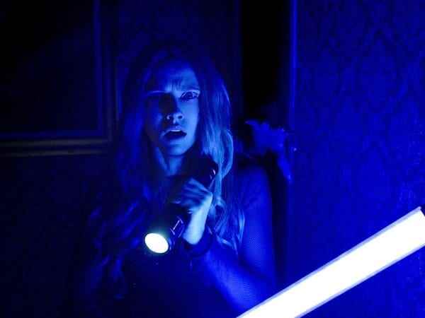 Ein Standbild aus dem Horrorfilm „Lights Out“, in dem eine Frau im UV-Licht steht und eine Taschenlampe hält, während sich eine Hand von einer Tür hinter ihr ausstreckt.