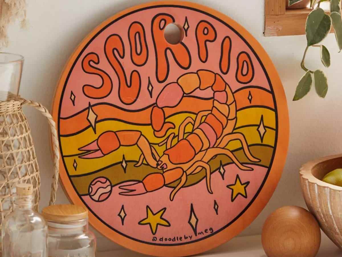 Ein buntes rundes Schneidebrett lehnt an der Wand auf einer Küchentheke.  Das Schneidebrett zeigt einen Skorpion und den Text „Skorpion“.