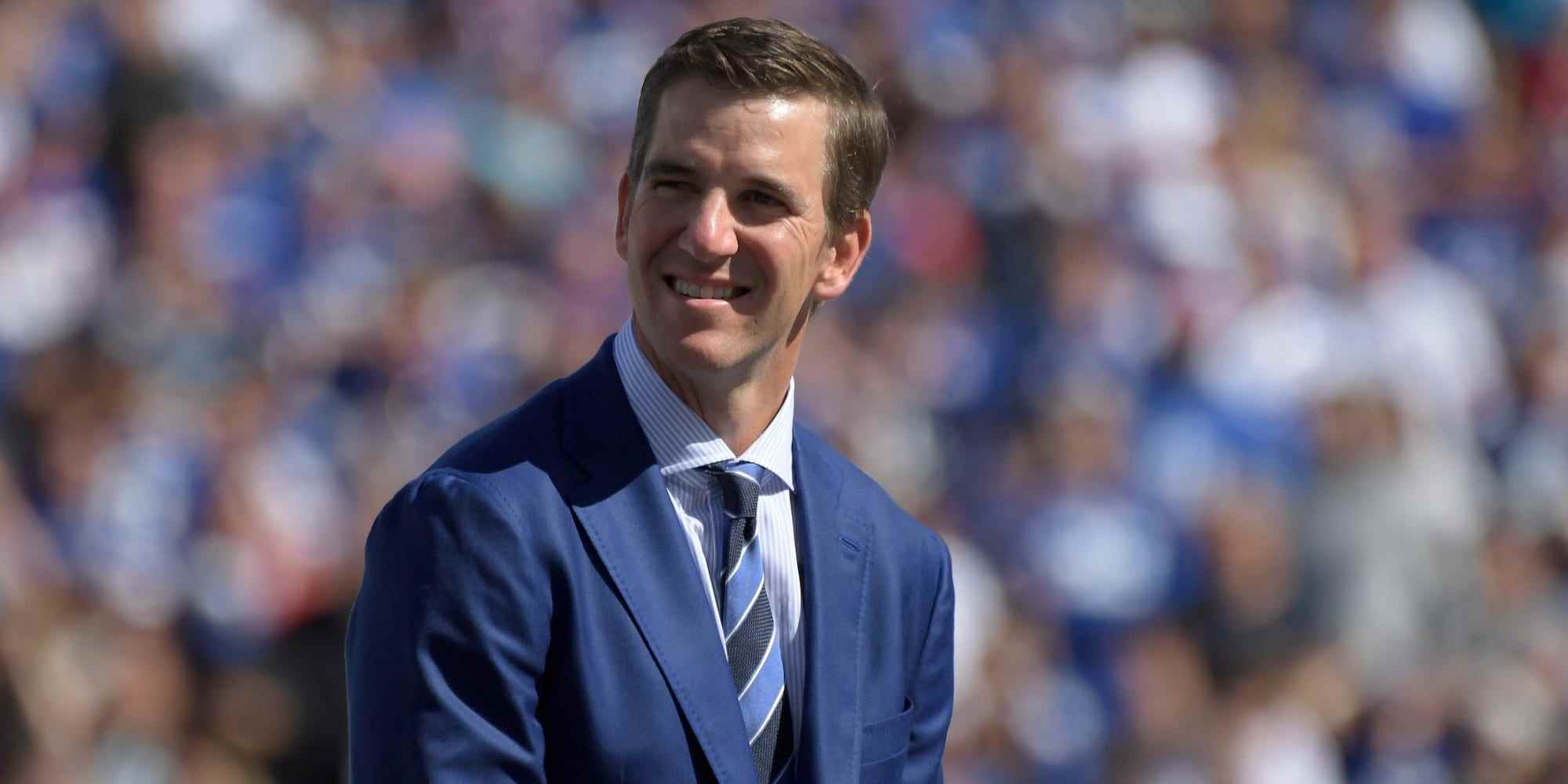 Eli Manning, gekleidet in einen Anzug, lächelt und schaut auf seine Ruhestandszeremonie im Giants-Trikot im Jahr 2021.