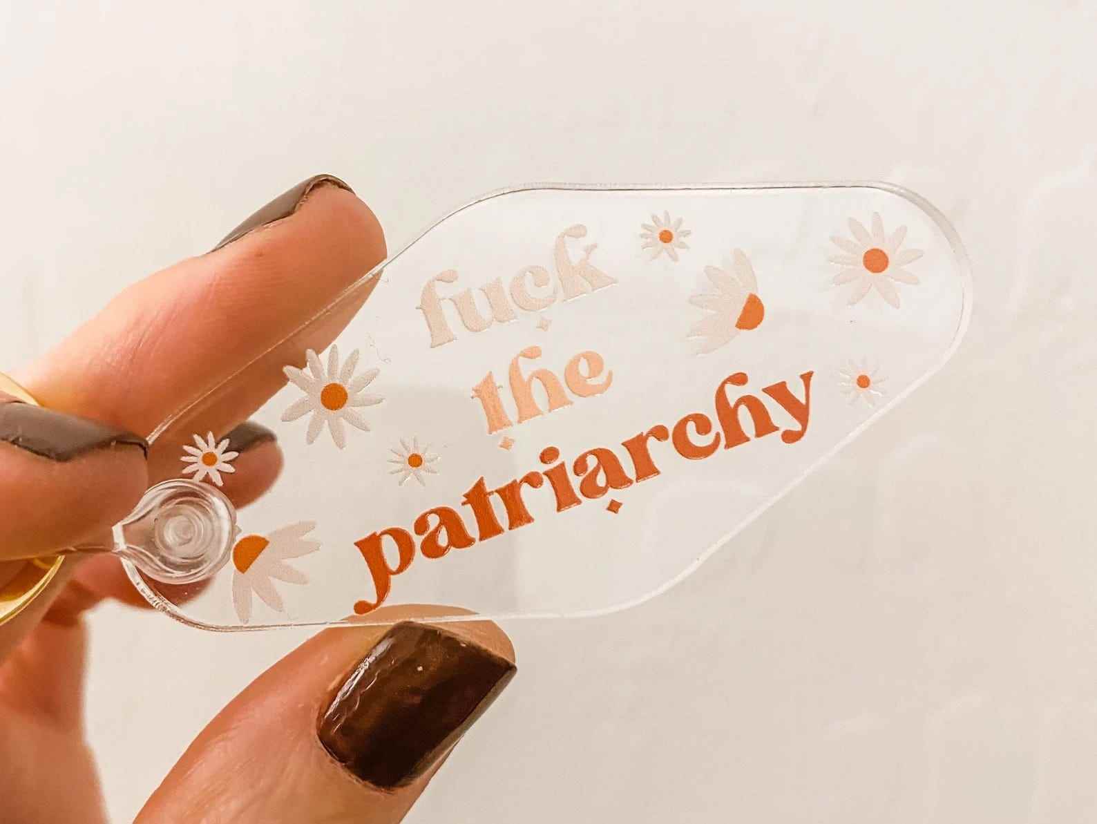 eine Hand, die einen durchsichtigen Schlüsselanhänger mit der Aufschrift „Fuck the Patriarchy“ hält.